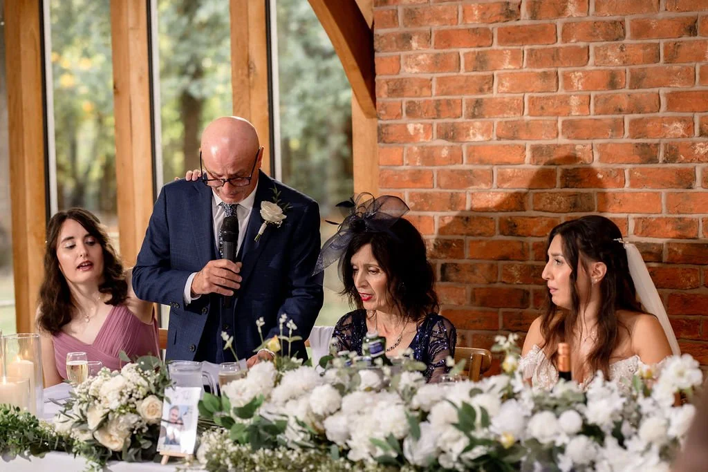 A wedding reception scene with a man in a suit giving a speech, two women in formal attire, and floral decorations on the table.
