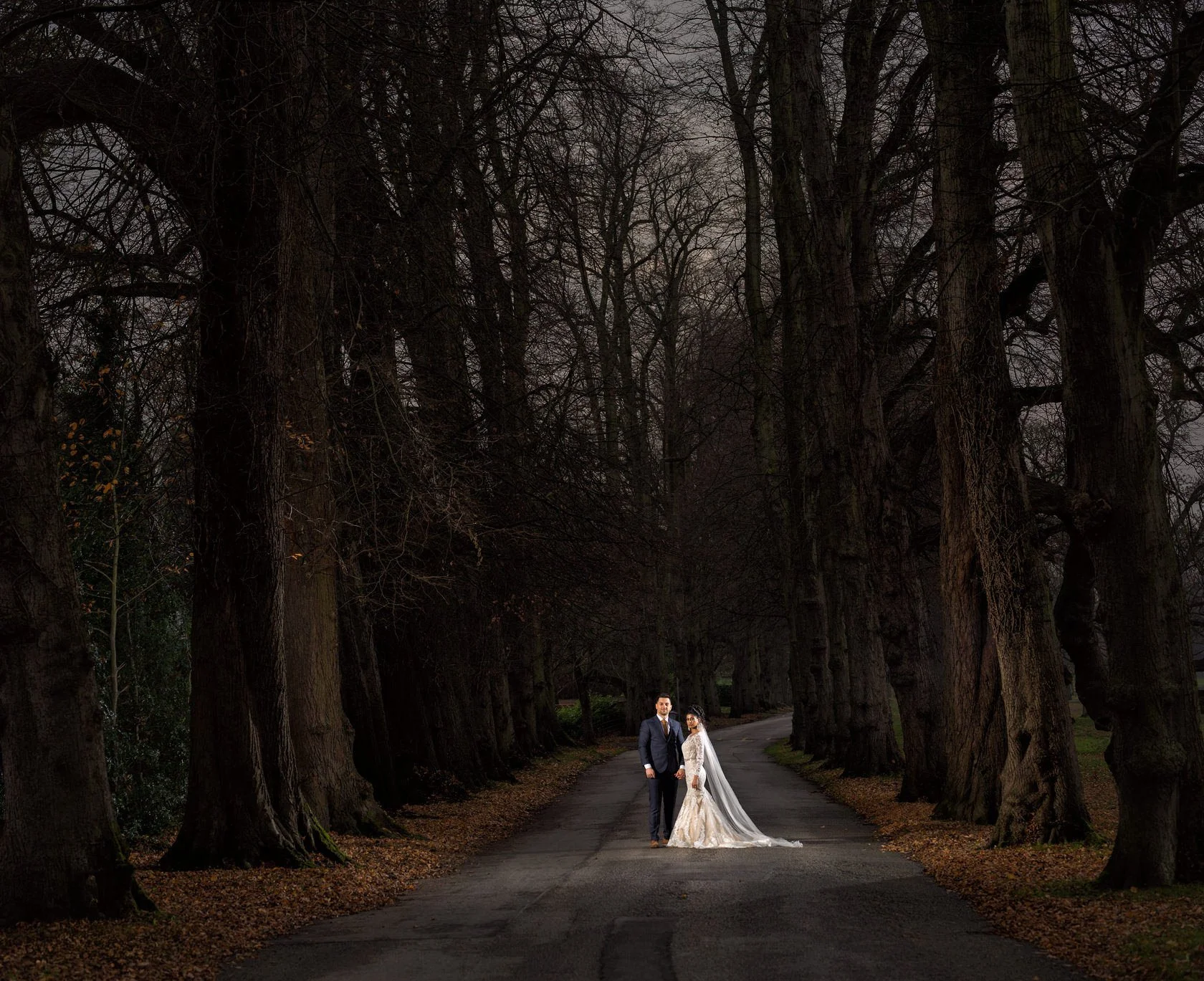 Bride and groom standing together on the driveway leading through a large area of tall trees during an Autumn evening.