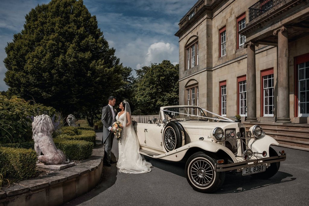 A newlywed couple stands beside a vintage white convertible car outside a grand stone building with columns, set in a well-manicured garden.