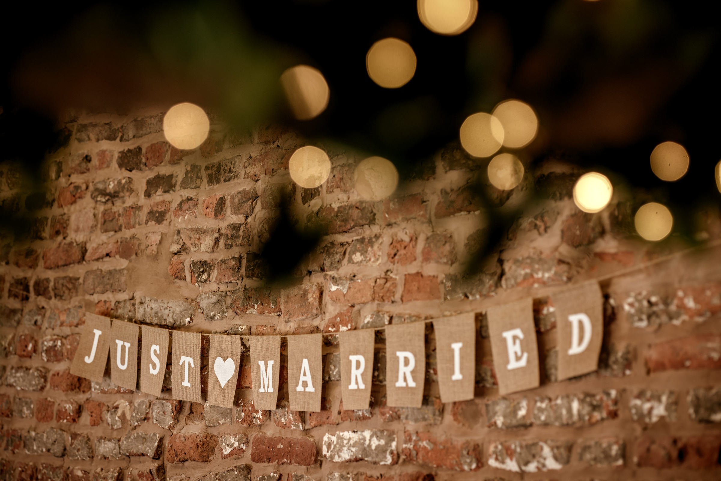 A 'Just Married' banner with brown paper flags and white letters hangs against a brick wall, decorated with soft warm string lights creating a festive atmosphere.