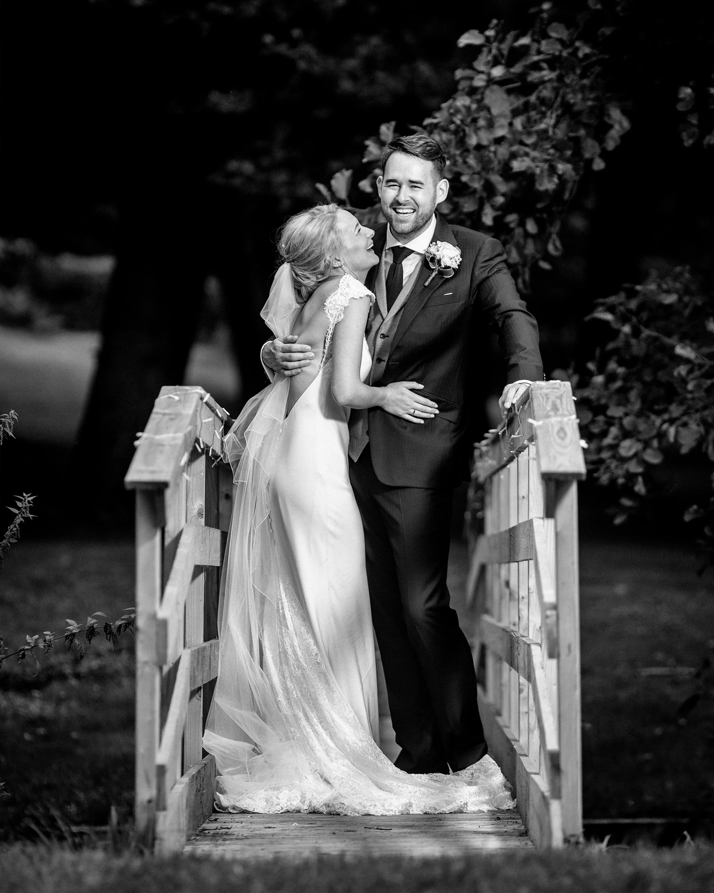 A bride and groom sharing a joyful moment on a small wooden bridge outdoors, black and white photograph.