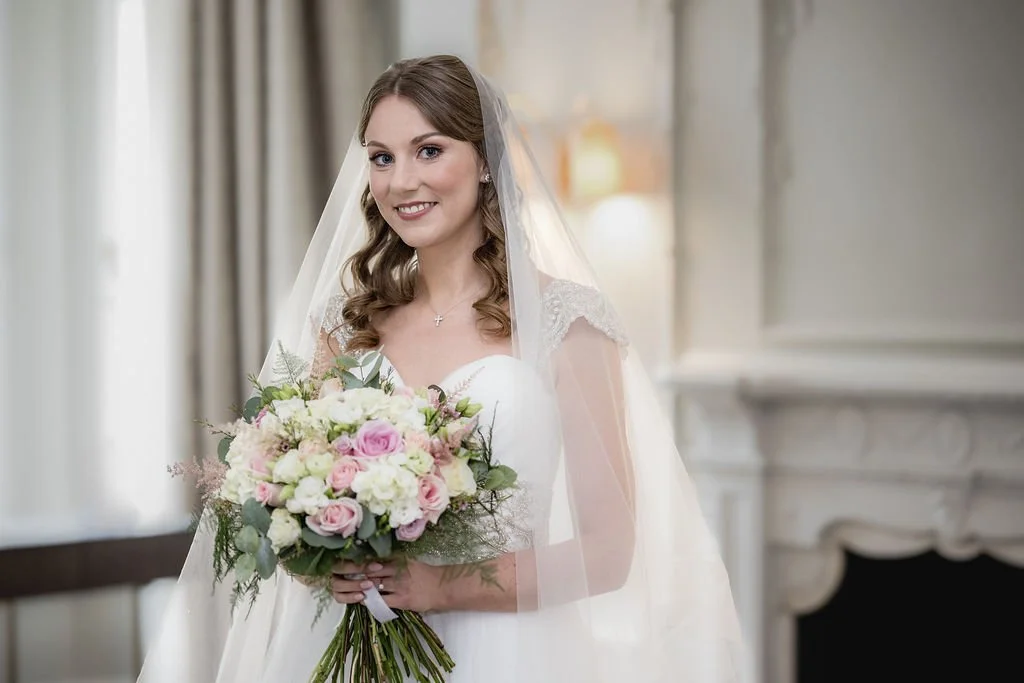 A bride in a wedding dress holding a bouquet of pink and white flowers, standing indoors near a fireplace.