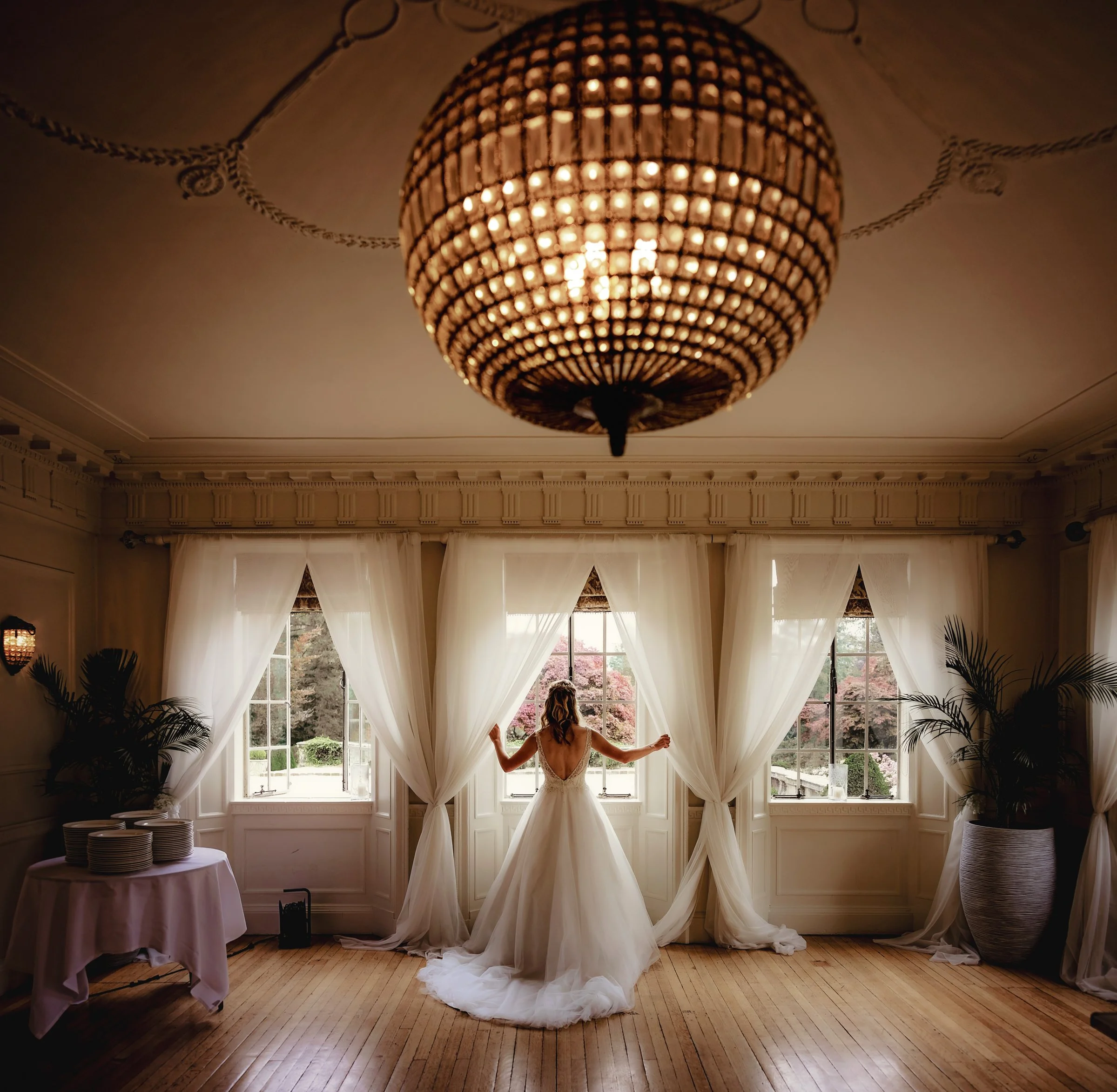 A woman in a white wedding dress standing in front of large windows with sheer curtains, holding the curtains open, in a decorated room with plants and a chandelier.