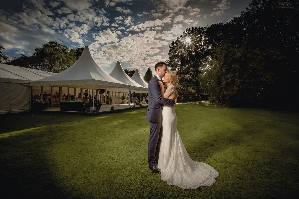 A bride and groom in wedding attire embrace outdoors on a grassy field with large white event tents and a partially cloudy sky in the background.