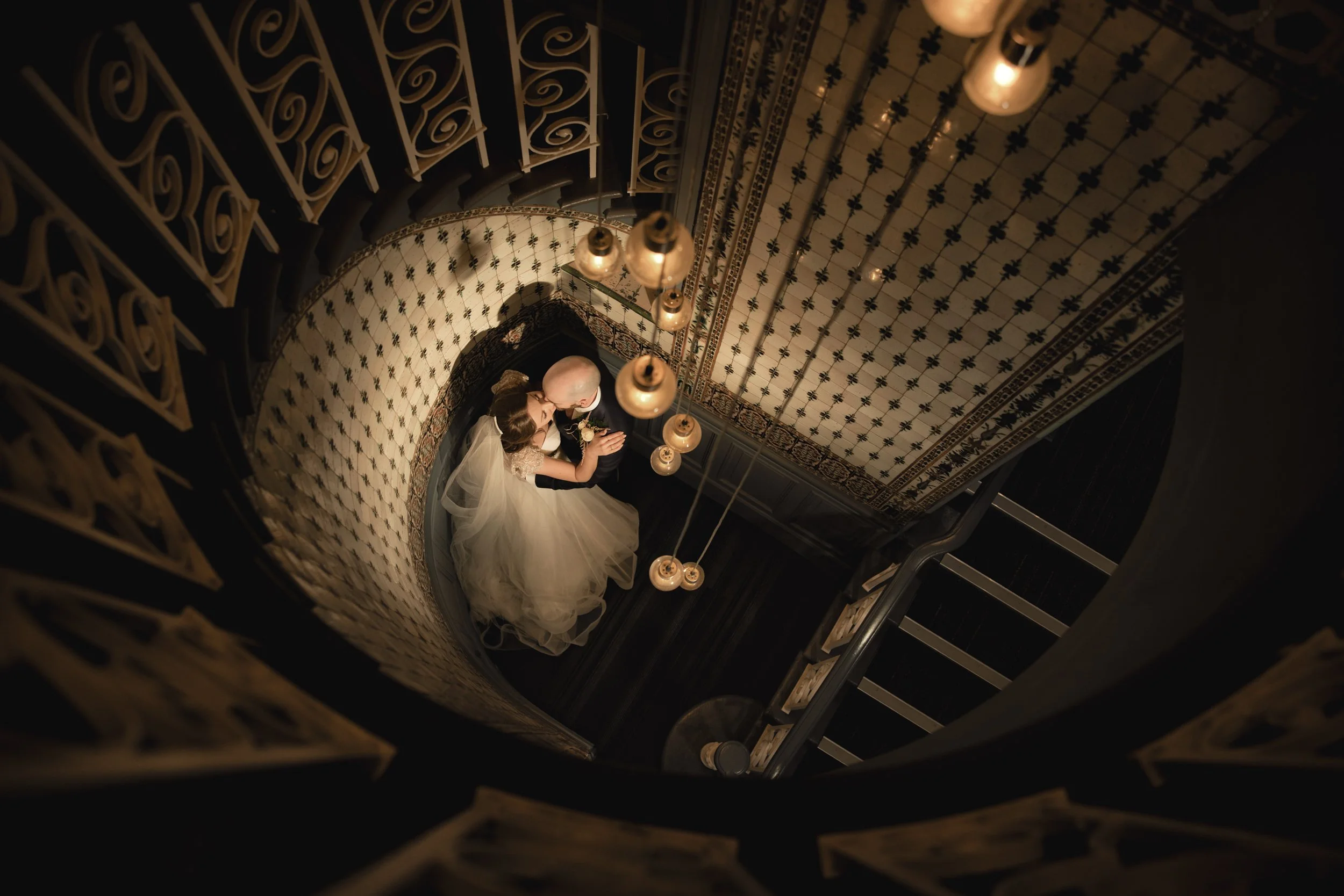 A bride and groom sharing a kiss on a staircase, photographed from above, with ornate decor and hanging lightbulbs.