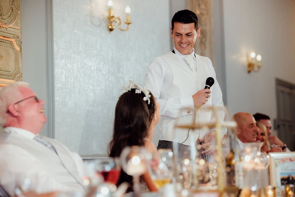 A man in a tuxedo giving a speech at a wedding reception, holding a microphone and smiling. Guests are seated at a table, laughing and listening.