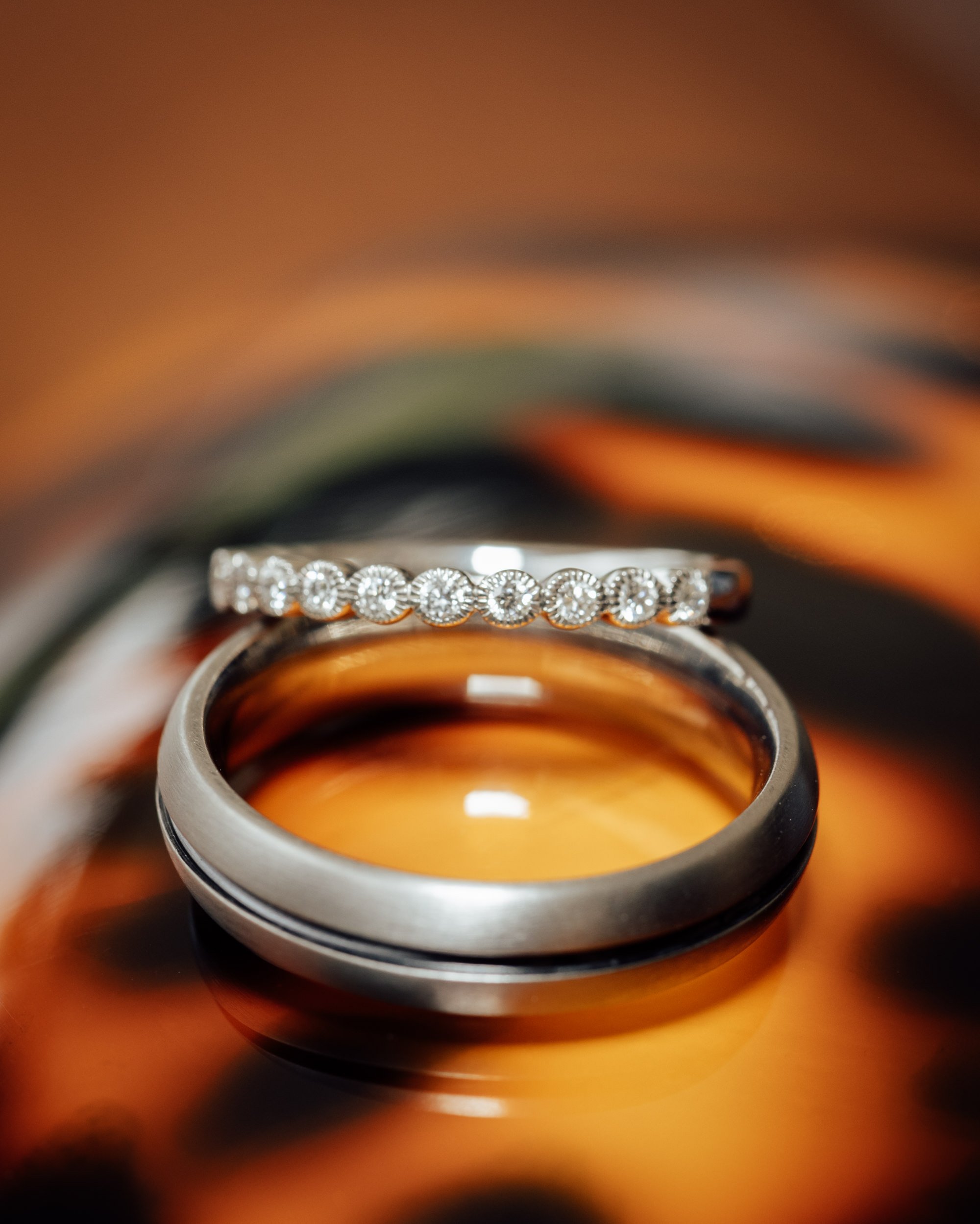 Close-up of a silver ring with small diamonds, placed inside a bottle with a brown-tan interior. The bottle is set on a reflective surface, giving a shiny background.