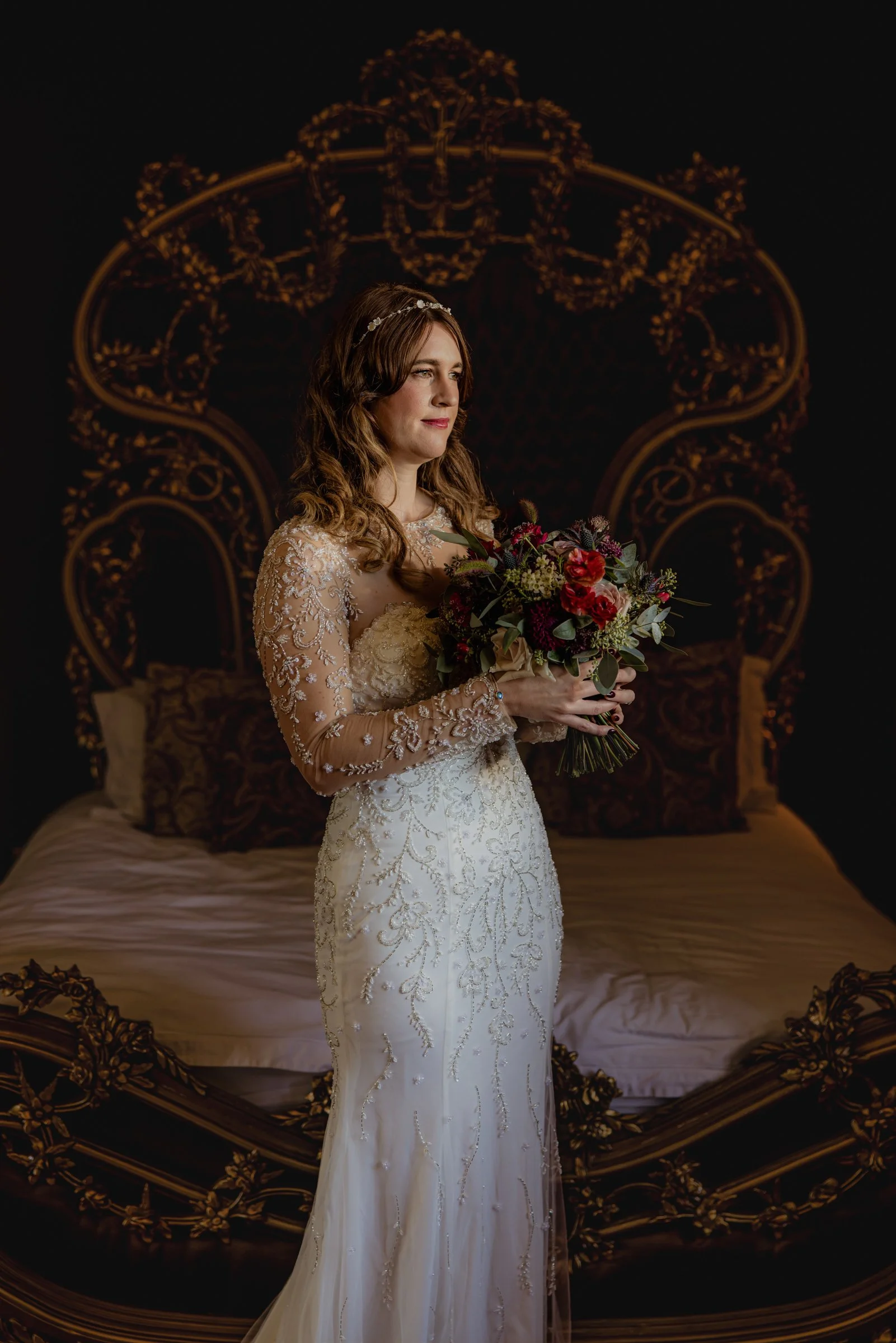 Bride in a detailed lace gown standing with a bouquet in front of a bed with an ornate headboard, dark background.