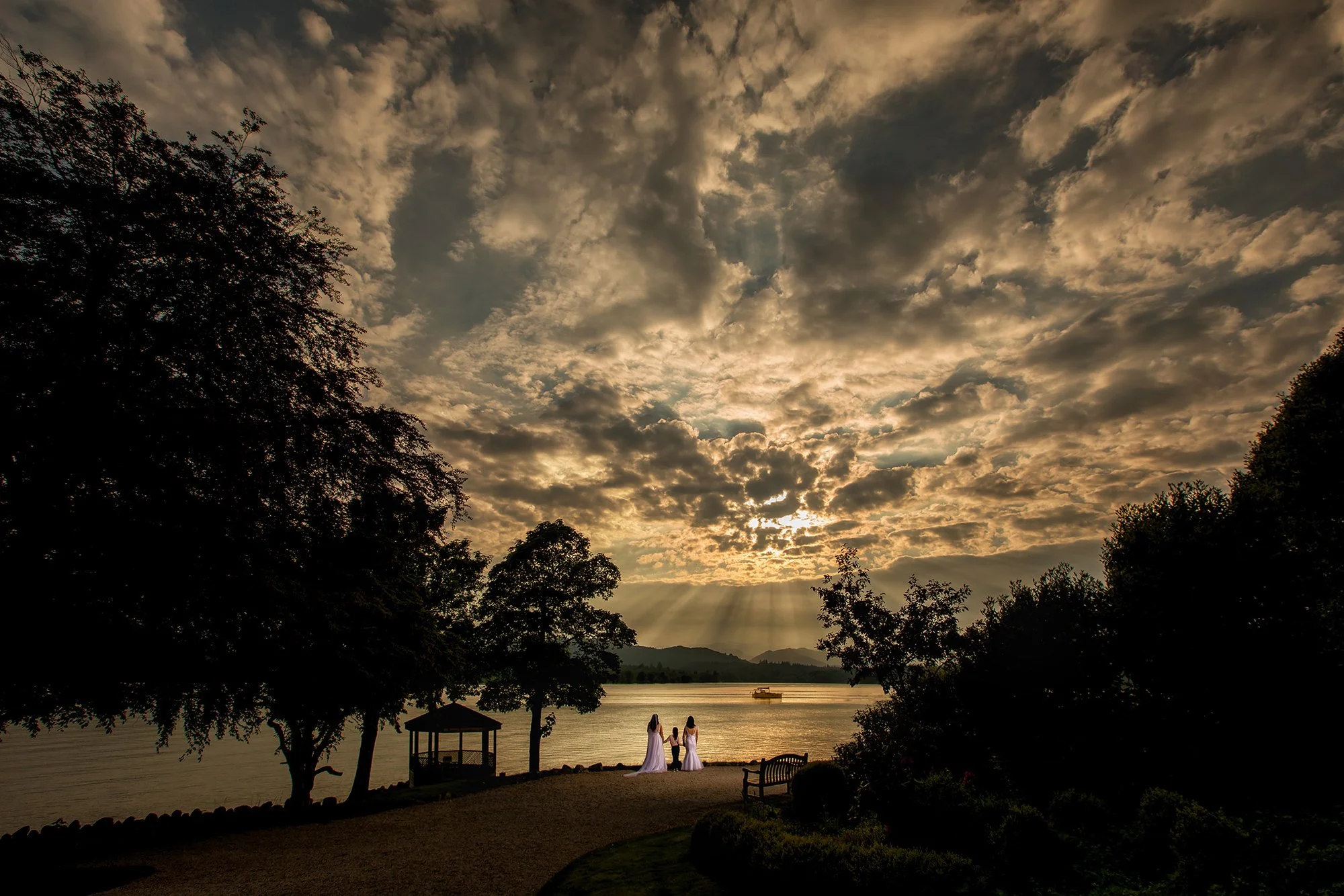 Silhouetted trees on a lakeside during sunset with two women in white dresses standing by the water, a small gazebo, and benches, with a partly cloudy sky and mountain range in the background.