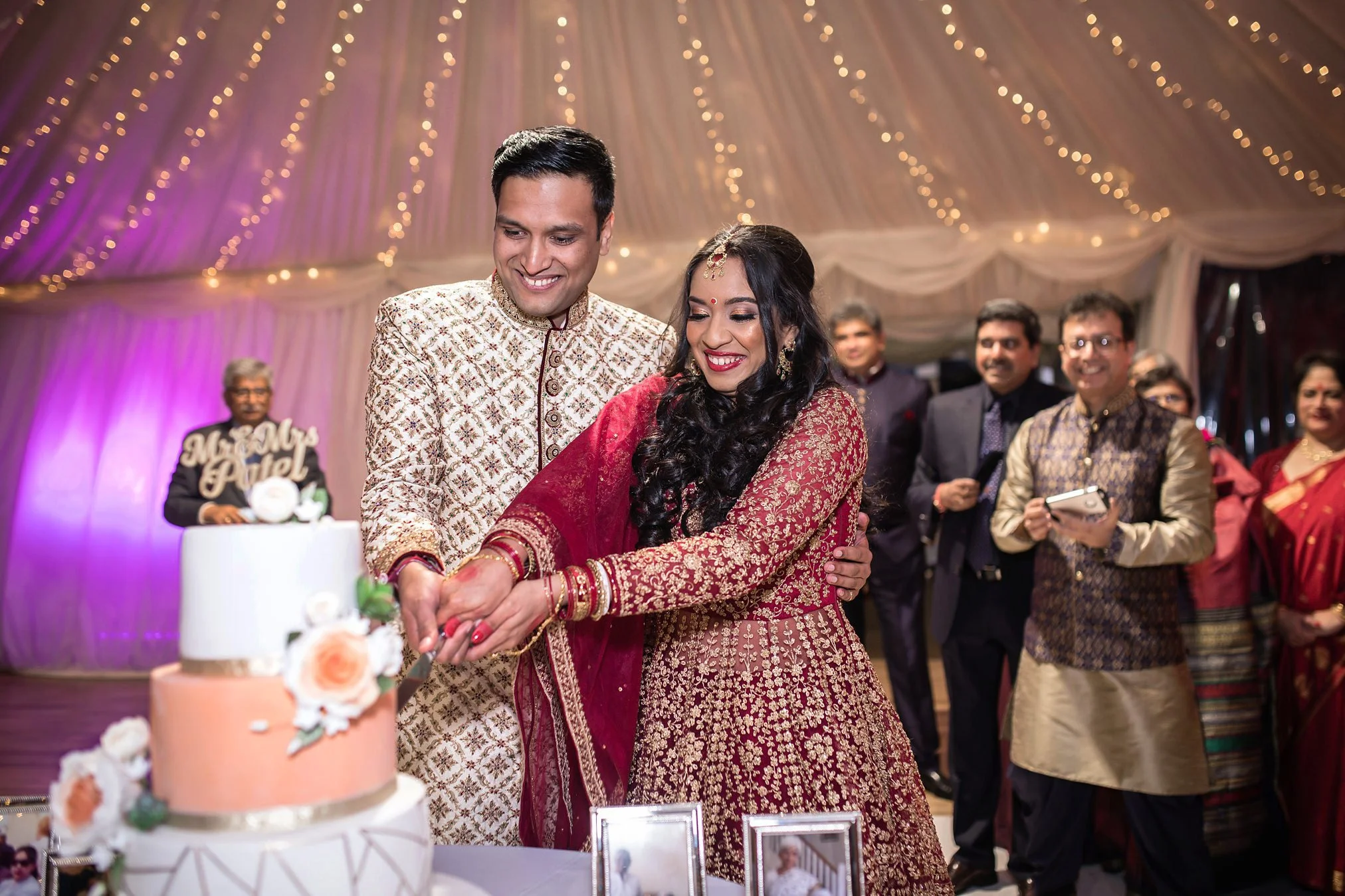 Asian bride and groom cut their wedding cake surrounded by smiling guests within a marquee at Thornton Manor.