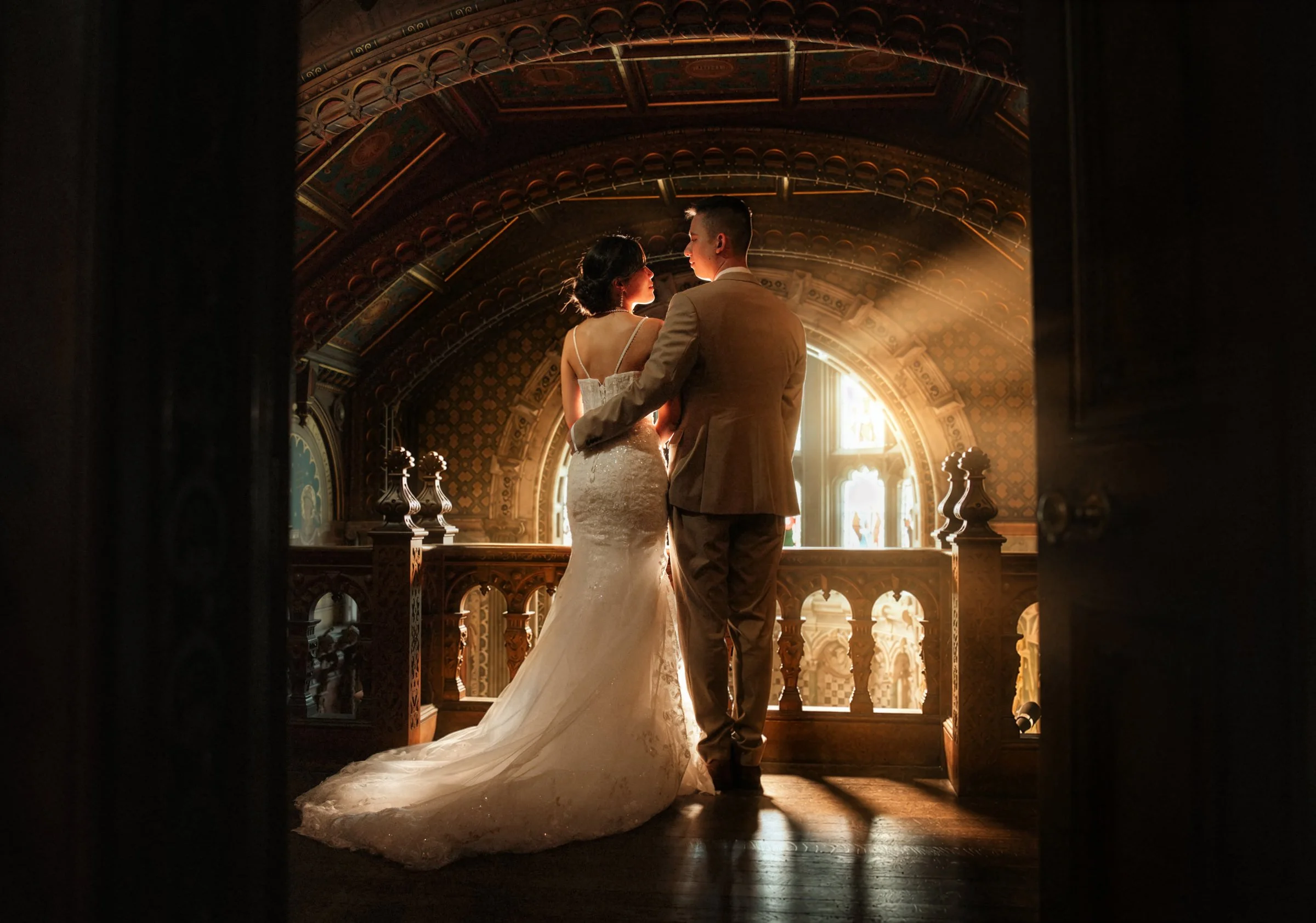 A bride and groom standing close together in a warmly lit, ornate room with intricate architectural details, viewed through a partially opened door.