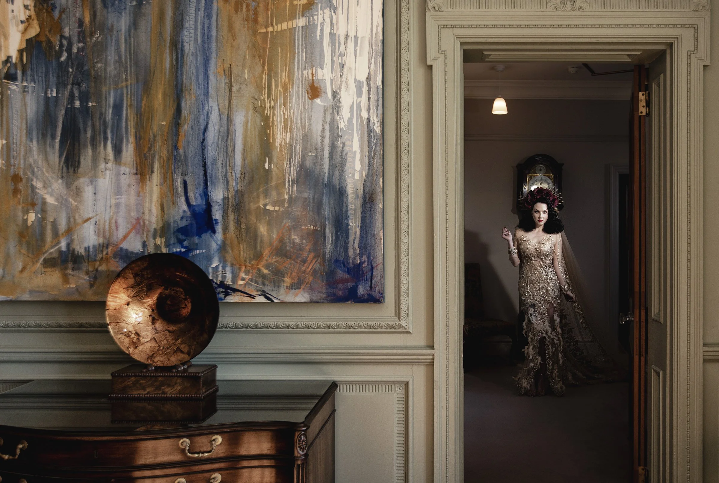 A woman in an ornate, gold and floral dress with a floral headpiece, standing in a doorway in a vintage-style room.