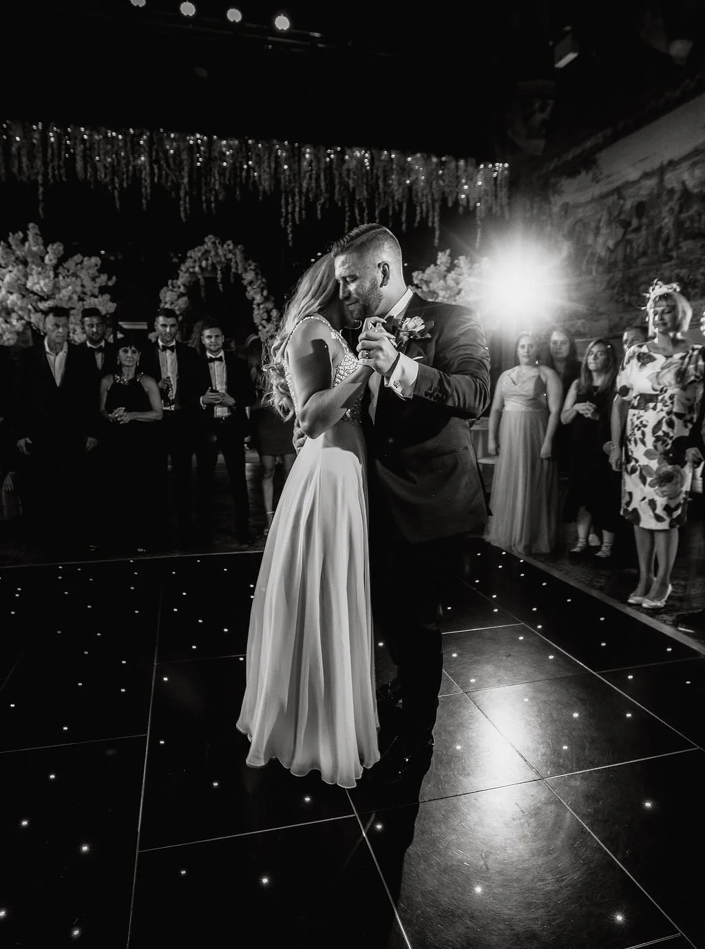 A black and white photo of a bride and groom sharing their first dance at a wedding reception, surrounded by guests and floral decorations.