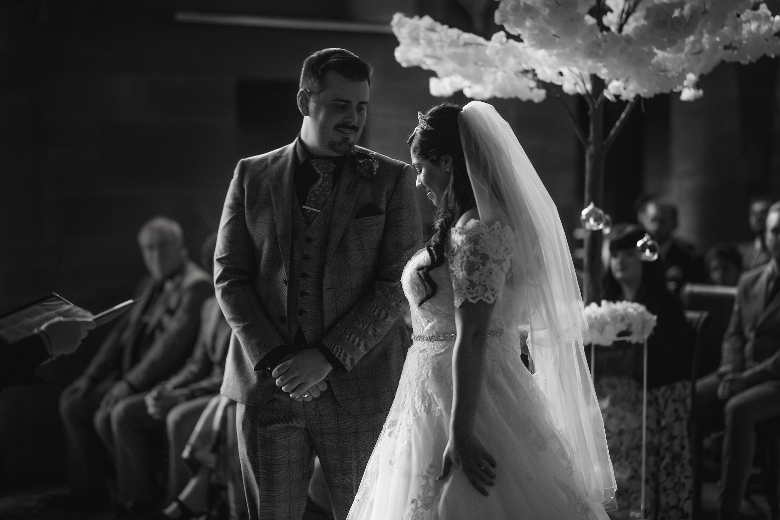 A black and white photo of a wedding ceremony with the bride and groom standing close, smiling quietly at each other, with guests seated in the background.