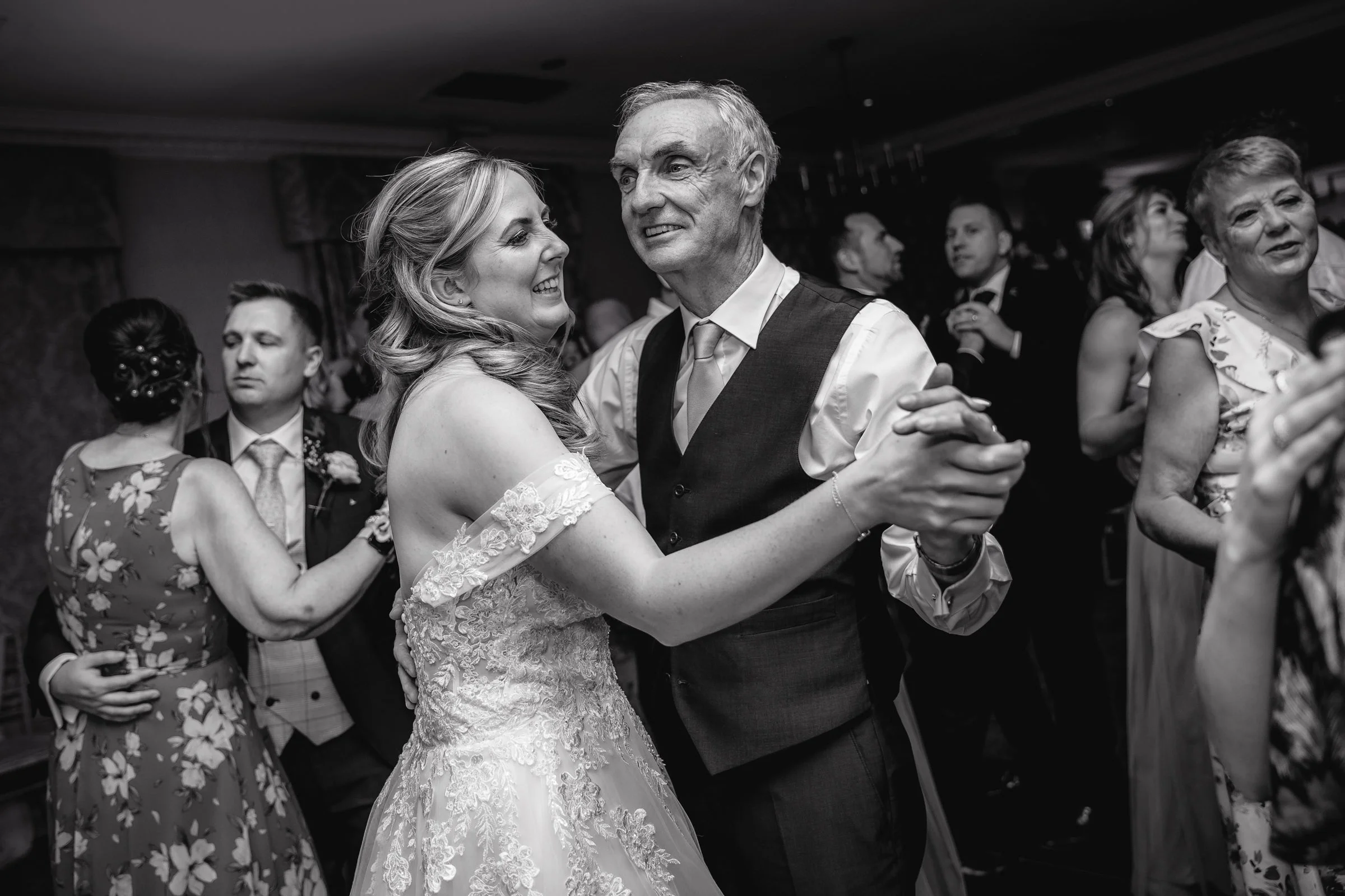 A bride and an older man dancing together at a wedding reception, smiling and looking at each other, surrounded by other guests dancing and socializing.