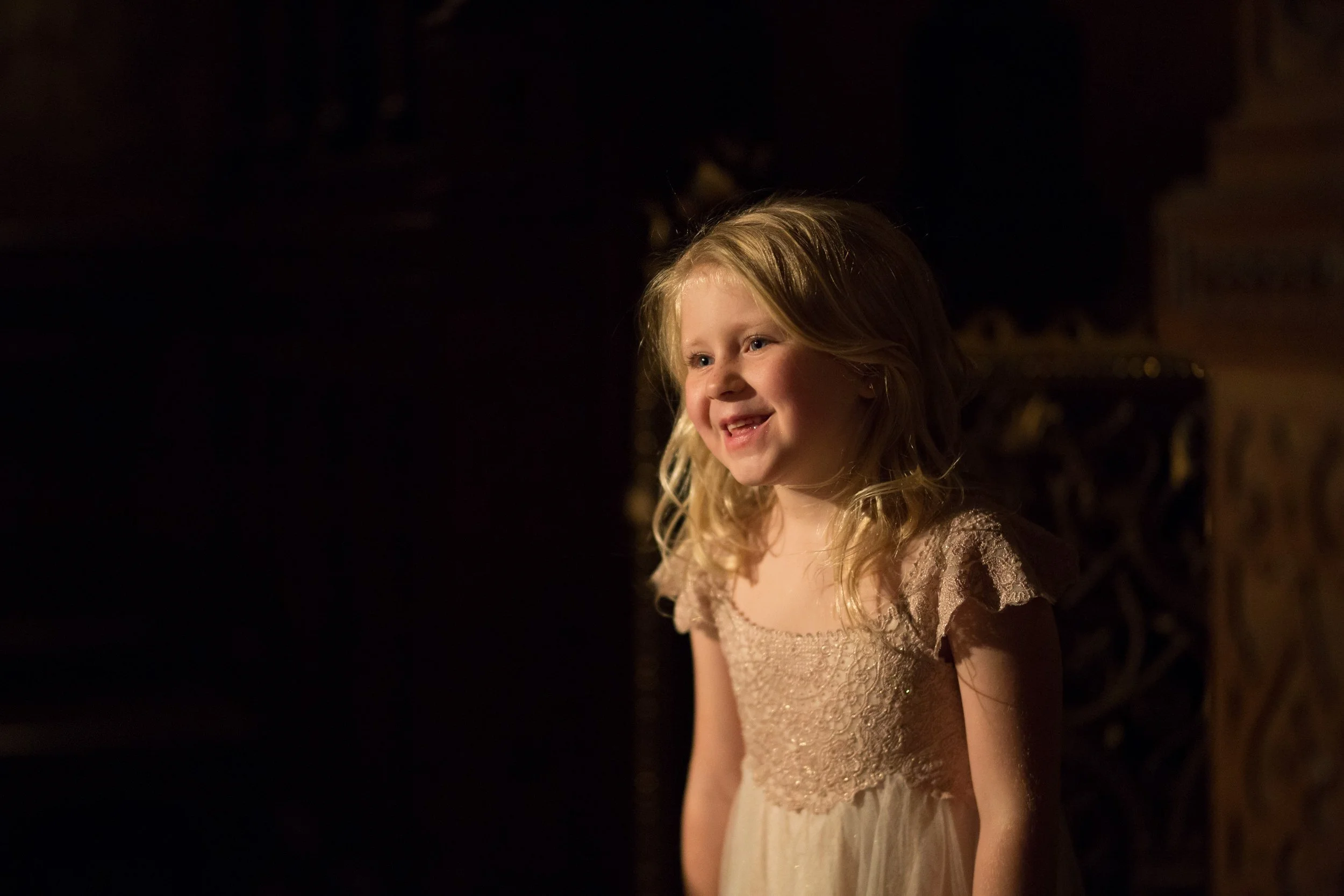 A young girl with blonde hair, wearing a light-colored dress, smiling in a dimly lit room with warm lighting.