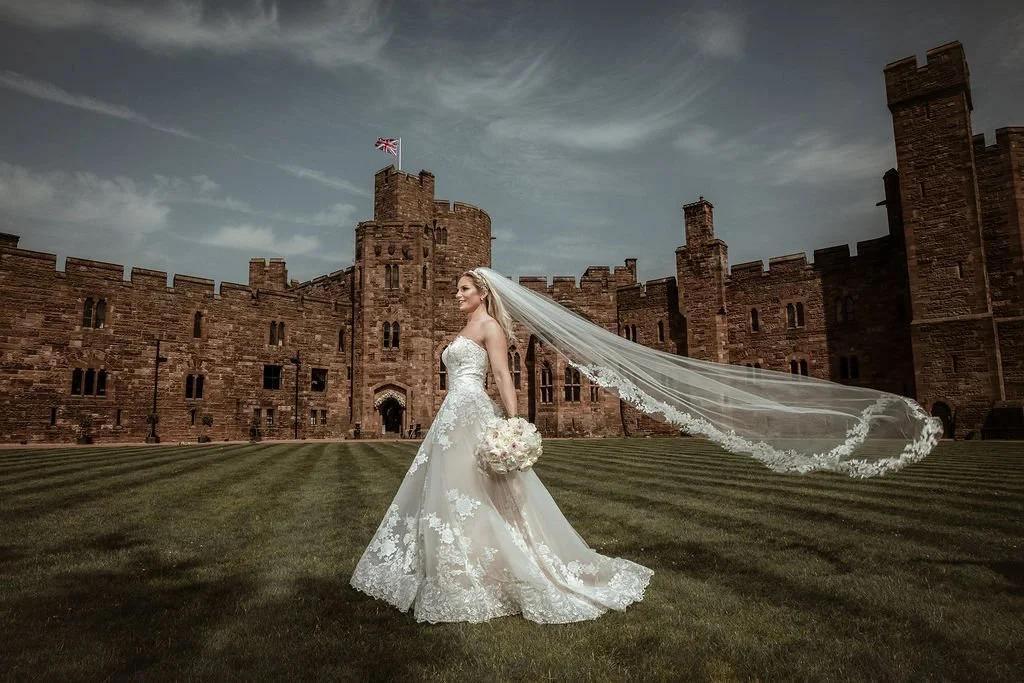 Bride in a white wedding dress holding a bouquet standing on grass in front of a historic castle with a flag on top, under a cloudy sky.