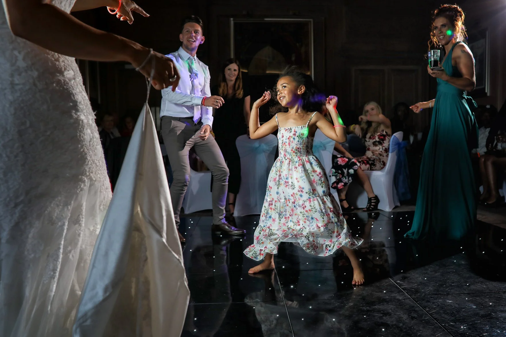 A young girl in a floral dress dancing on a black reflective floor at a celebration, surrounded by adults watching and enjoying the moment.