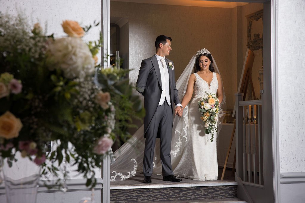 A bride and groom holding hands, standing inside near the door of a wedding venue, smiling, with the bride holding a bouquet of flowers and wearing a lace wedding gown with a veil, while the groom wears a gray suit with a tie.