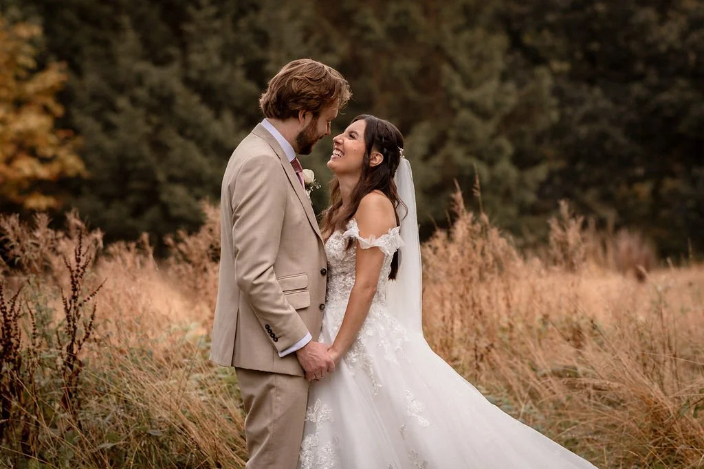 A bride and groom on their wedding day standing in a field of tall grass with trees in the background, smiling and holding hands.
