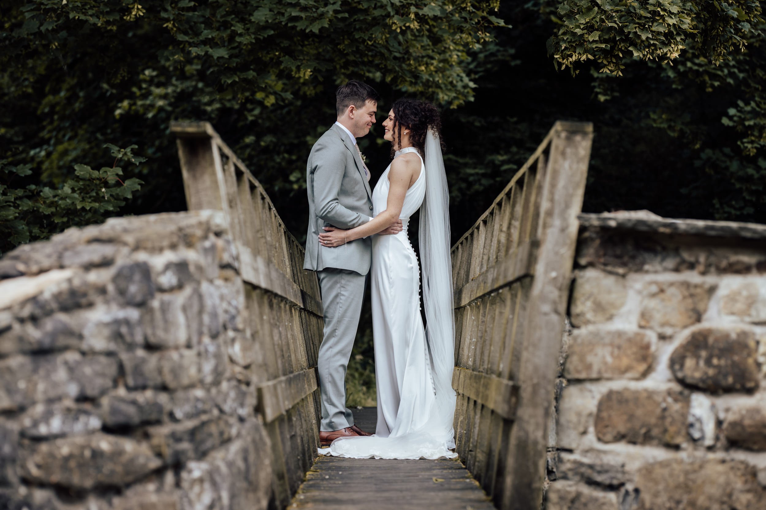 A bride and groom standing close together on a wooden bridge surrounded by lush green trees, sharing a tender moment on their wedding day.