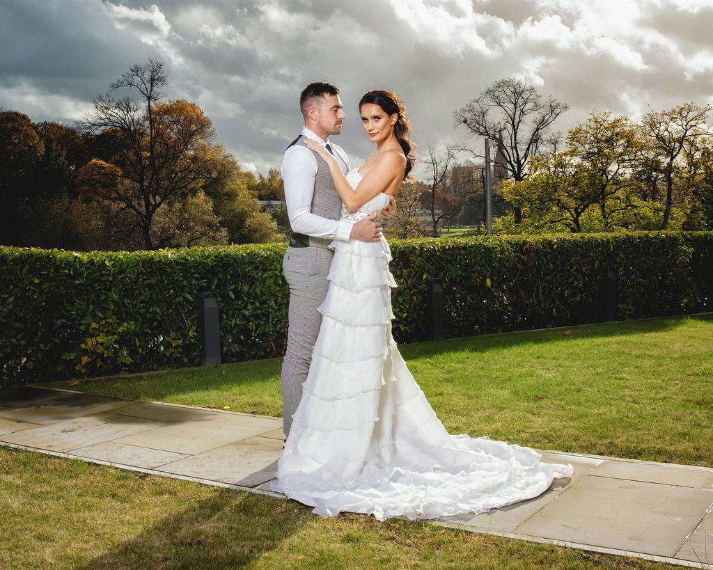 A newlywed couple posing outdoors on a paved walkway, with a lush green hedge and trees in the background, under cloudy skies.