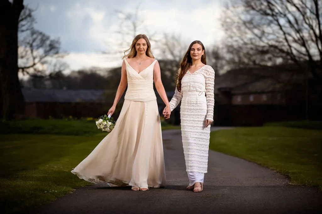 Two women in wedding dresses holding hands and walking on a pathway outdoors, with trees and a cloudy sky in the background.