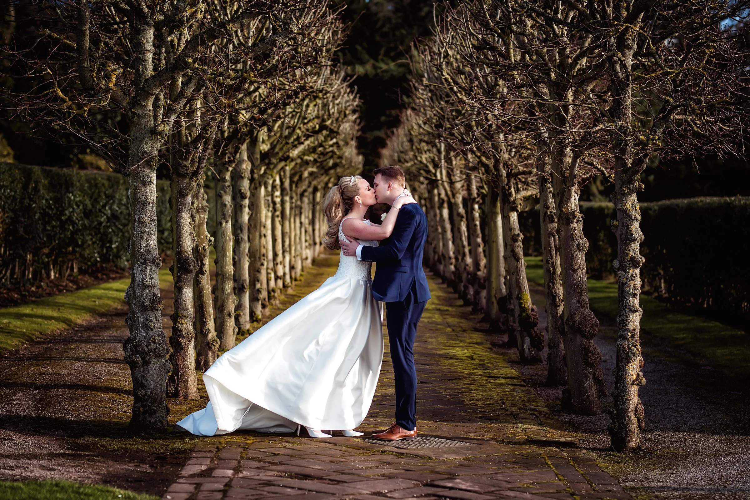 A bride and groom sharing a kiss in a tree-lined pathway, bride in a white wedding gown and groom in a navy suit.