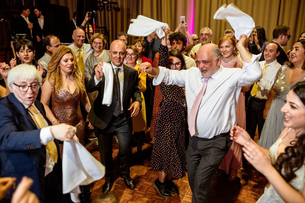 People dancing and celebrating at a party or wedding reception, with some holding napkins or cloths in the air, smiling and having a good time.