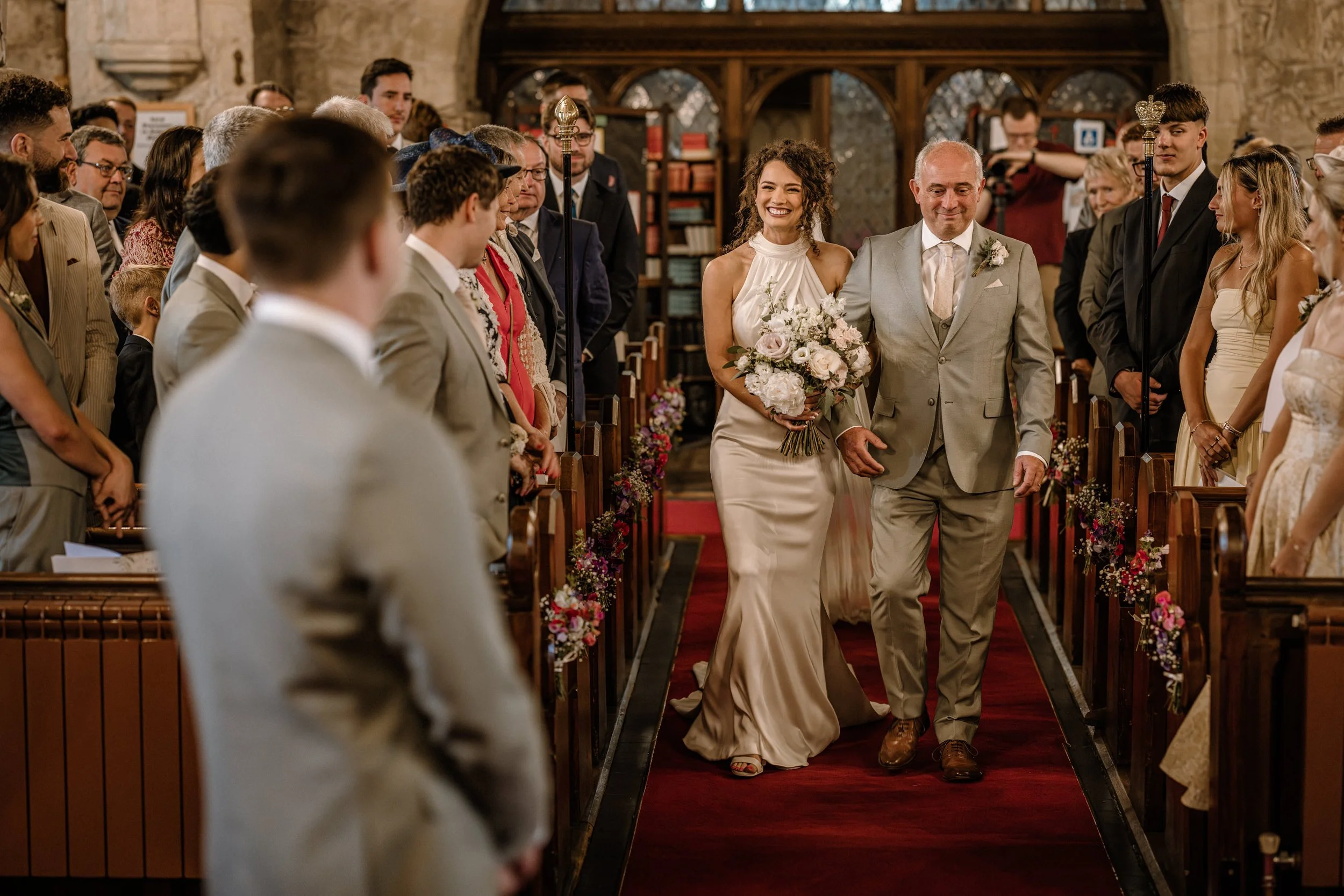 A woman in a white dress smiling as she walks down the aisle at her wedding, accompanied by a man in a light-colored suit.