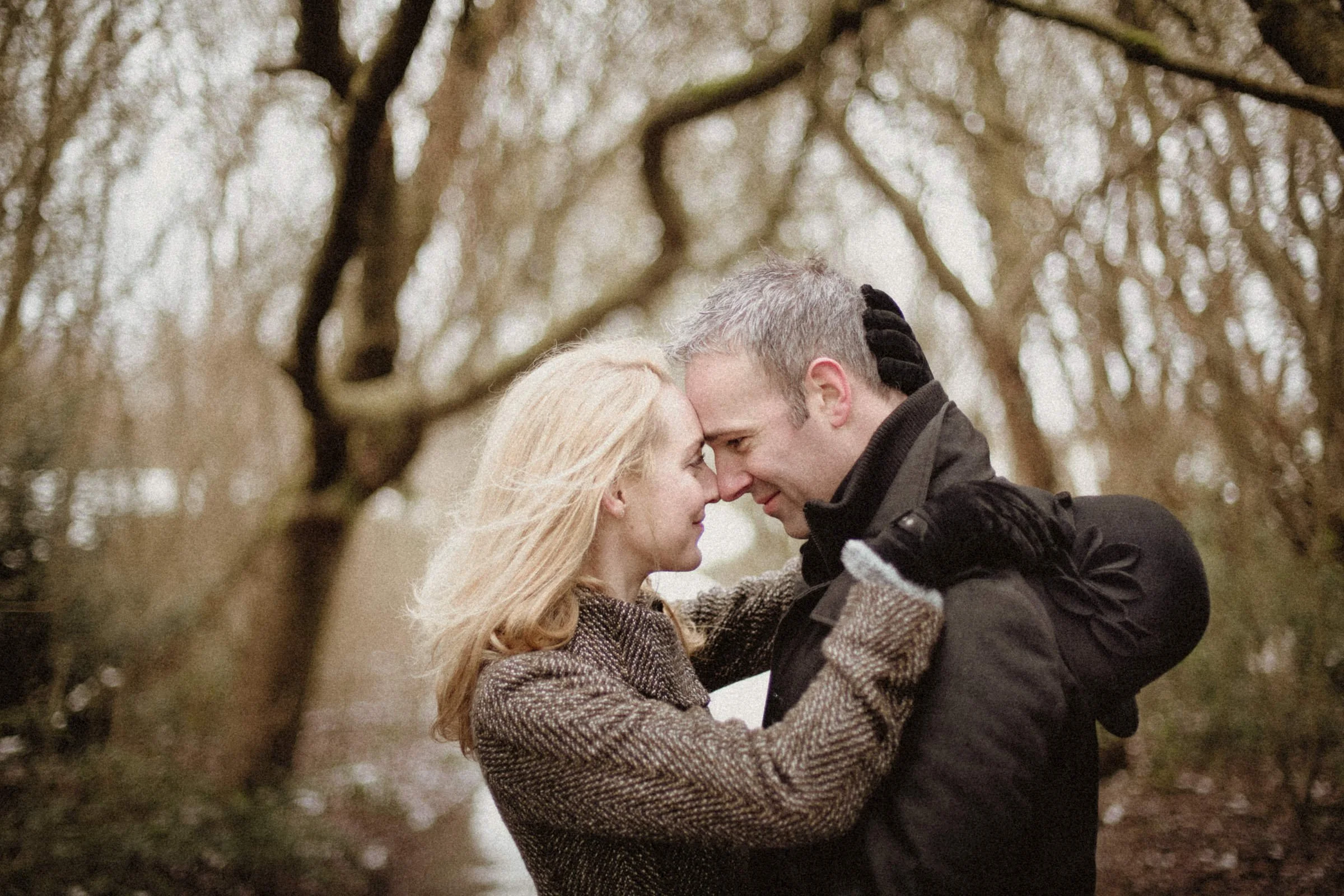 A man and woman facing each other closely, touching foreheads, outdoors in a wooded area with bare trees, on a cool day.