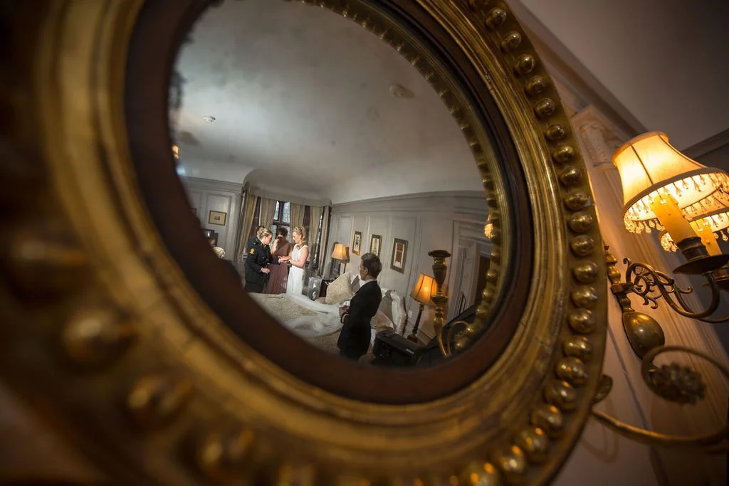 Reflection in an ornate gold-framed mirror shows a hotel room with four people getting ready, surrounded by lamps, curtains, and framed pictures.