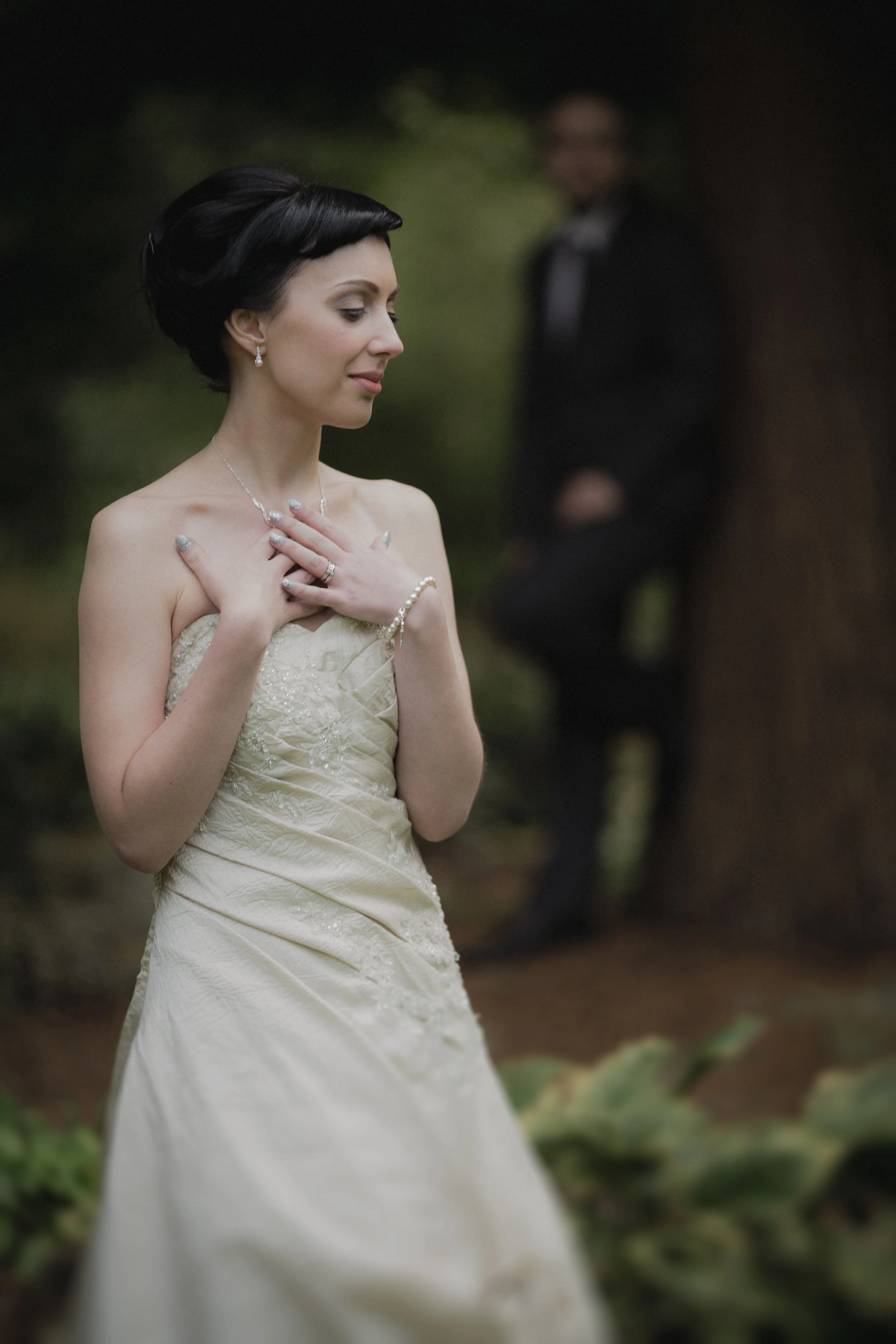 A bride in a strapless wedding dress with her hands on her chest, standing outdoors with a blurred groom in the background.