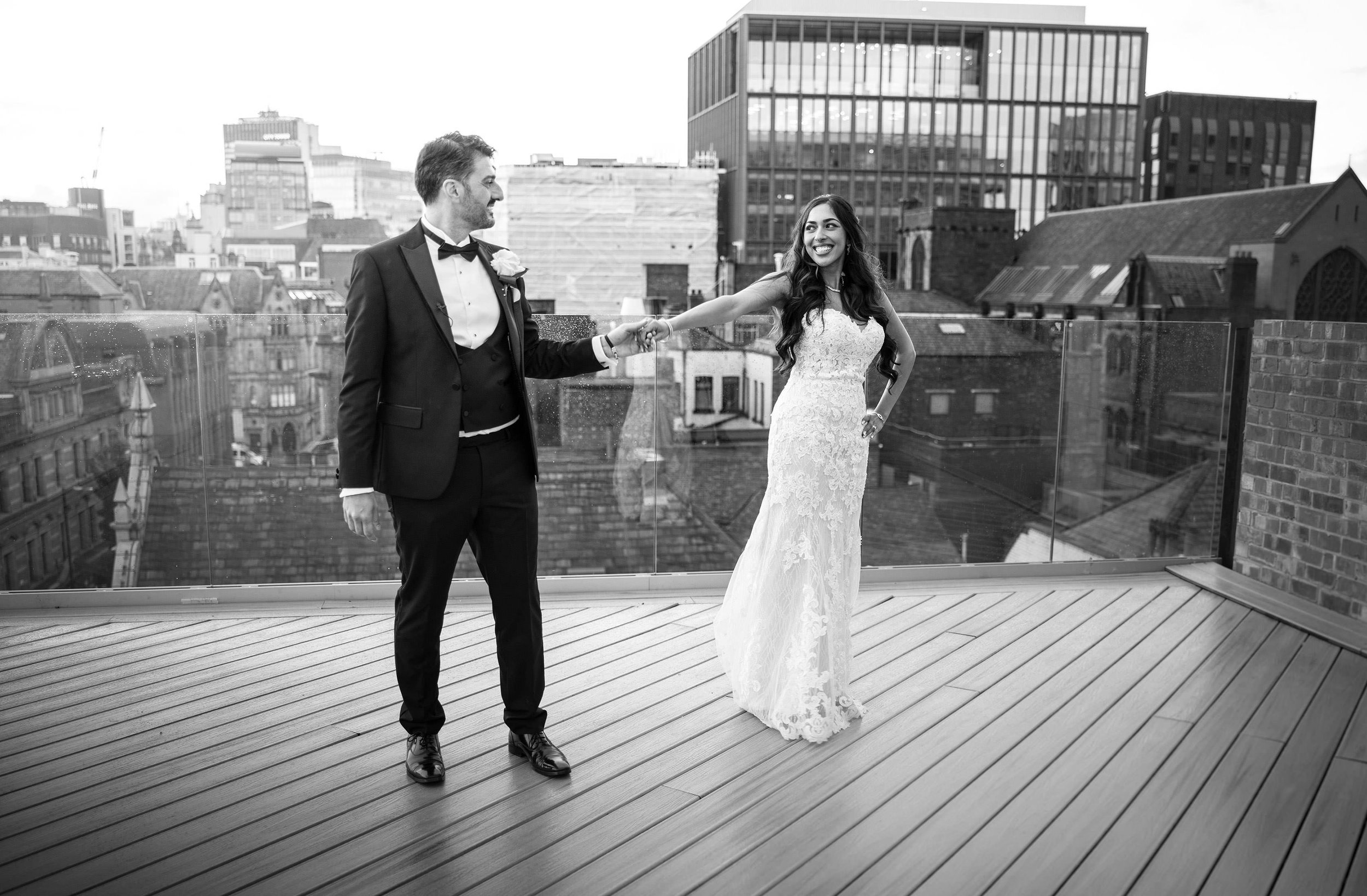 A man in a tuxedo with a boutonniere and a woman in a lace wedding gown on a rooftop with a city skyline in the background, holding hands and smiling at each other.