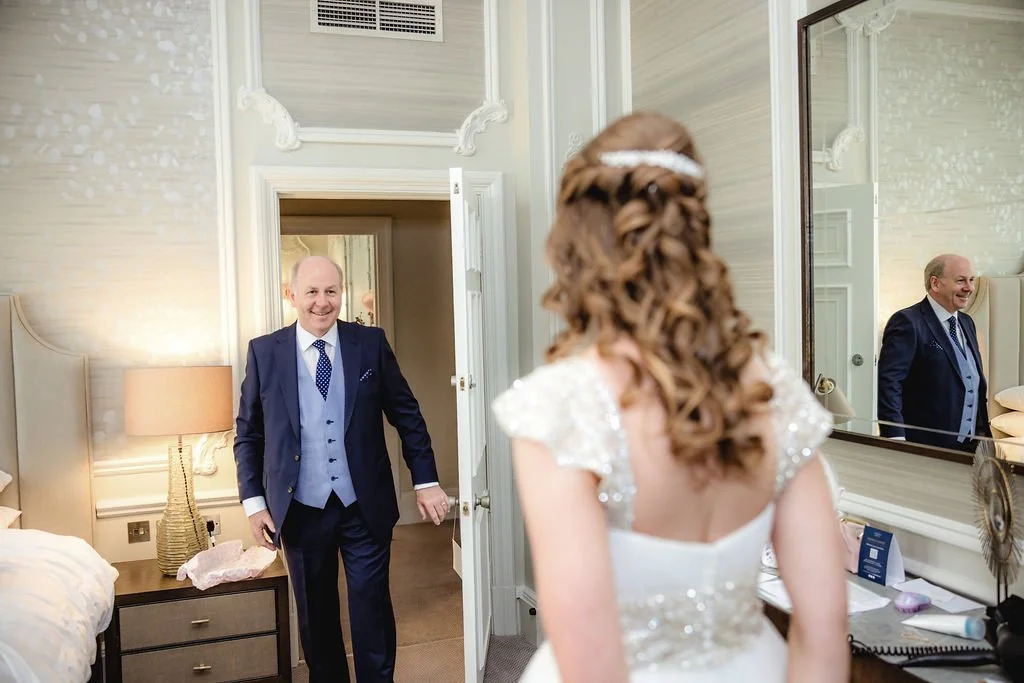 A man in a navy blue suit and a woman in a wedding dress stand in a hotel room, smiling at each other. The man is entering through a door, and the woman's back is to the camera. The room has a bed, a lamp, and a large mirror.