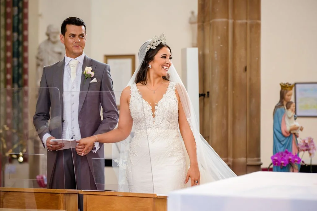 A bride and groom standing together inside a church, holding hands. The bride is wearing a white wedding dress and veil, and smiling. The groom is dressed in a gray suit with a white shirt and tie, holding a piece of paper, looking at the bride.