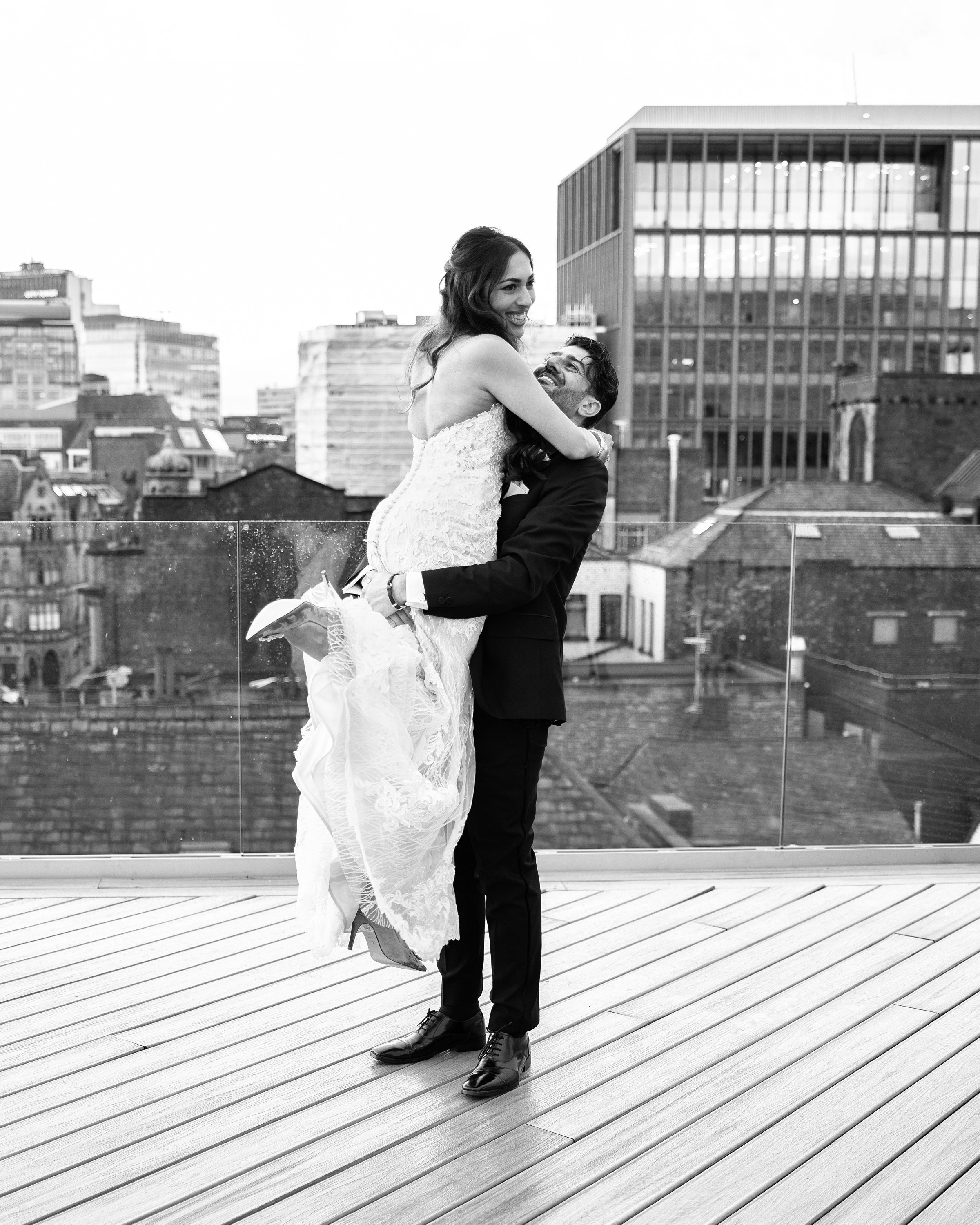 A man in a suit lifting a woman in a wedding dress on a rooftop with city buildings in the background, black and white photo.