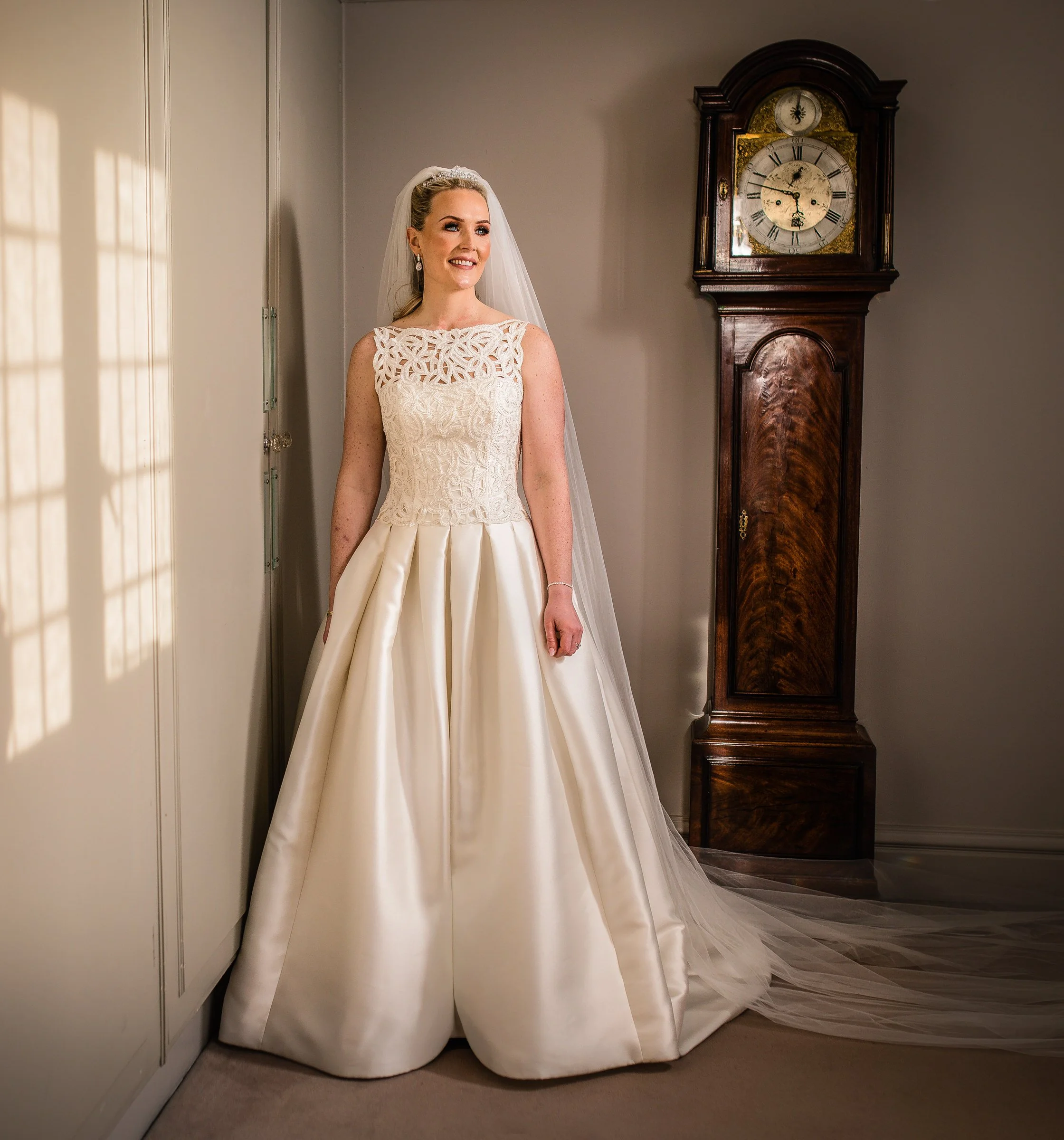 A bride in a white wedding gown and veil standing indoors near a wall and next to a tall wooden grandfather clock.