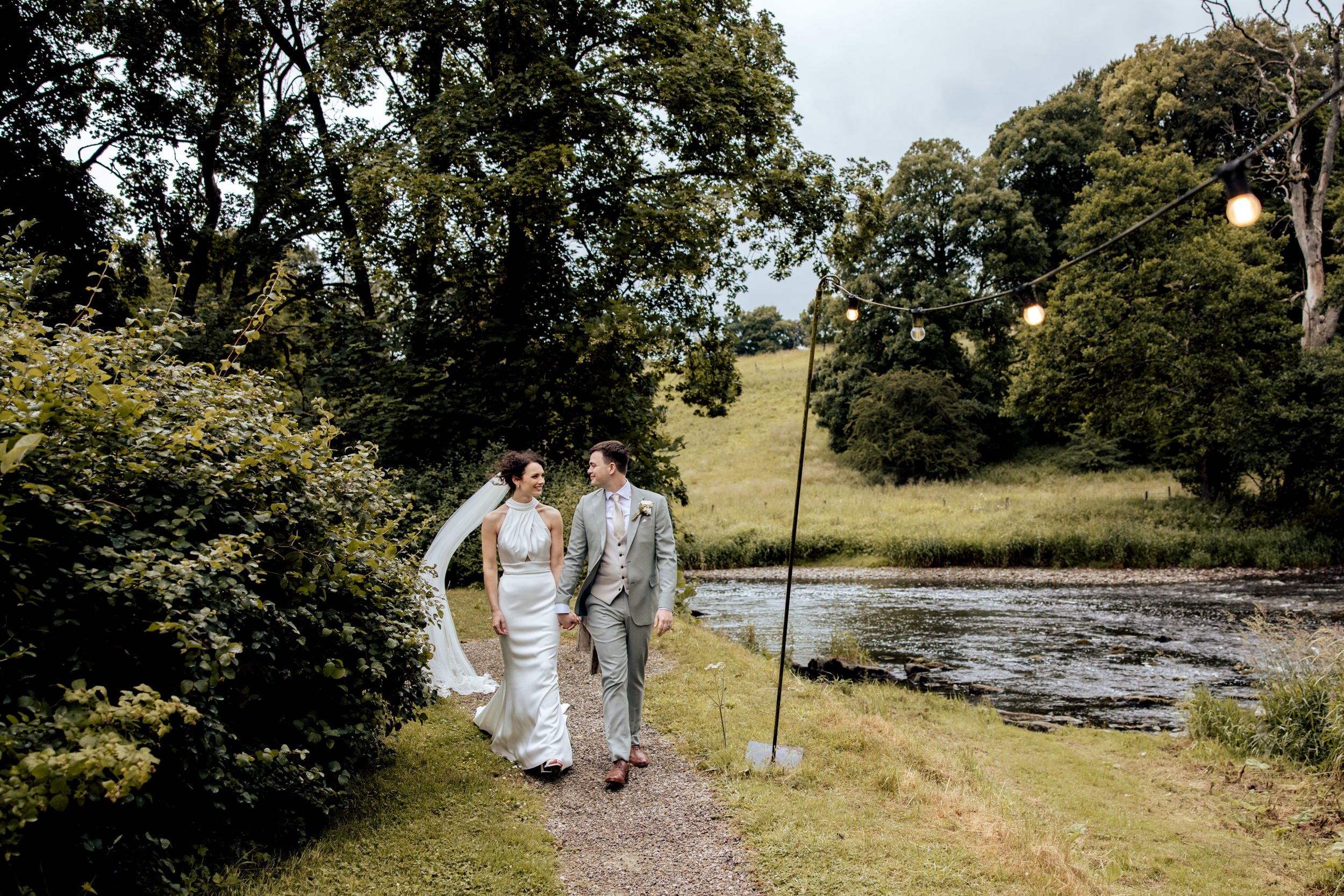 A bride and groom holding hands and walking along a grassy path beside a river during their outdoor wedding. The bride is in a white wedding dress, and the groom is in a light gray suit. Overhead string lights are hung among the trees, and a forested