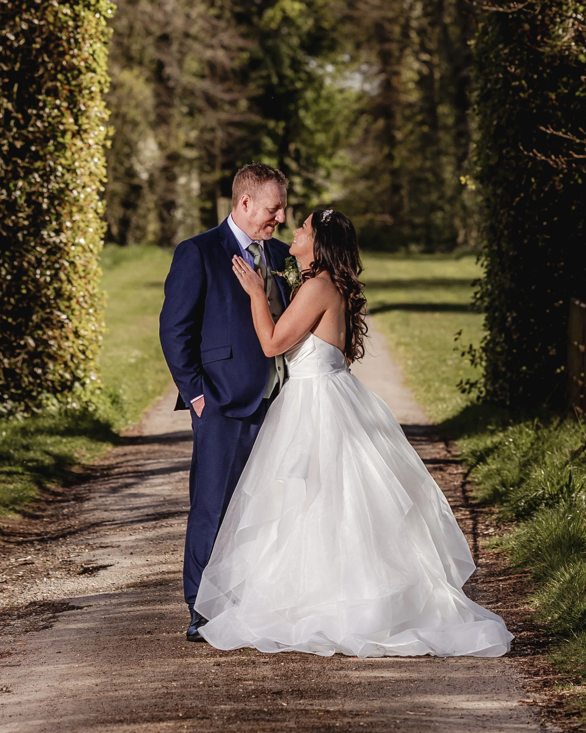 A bride in a white wedding gown and a groom in a dark blue suit stand close together on a dirt path surrounded by greenery, sharing a tender moment.
