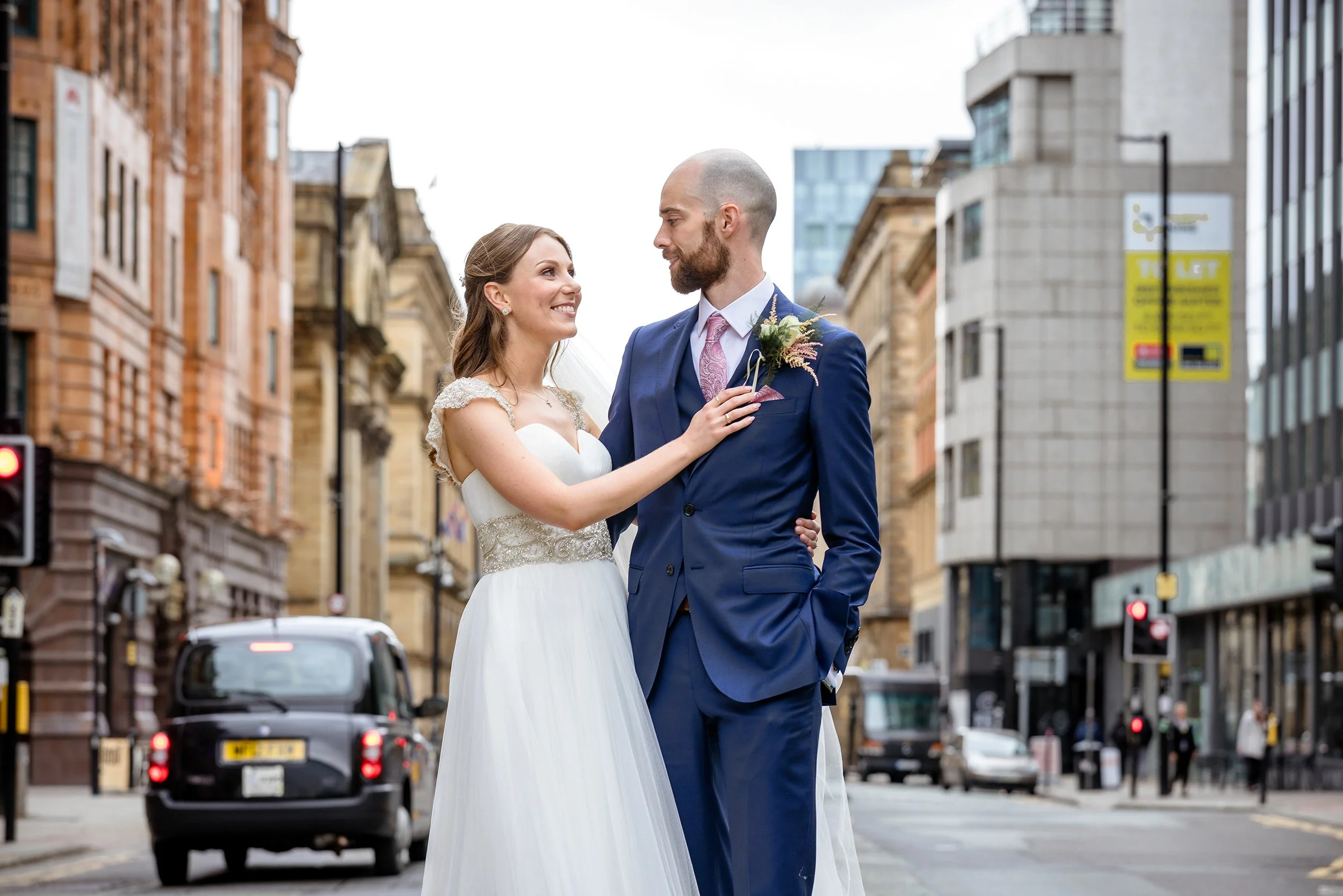 A bride and groom standing close together on a city street, smiling at each other, with tall buildings and cars in the background.
