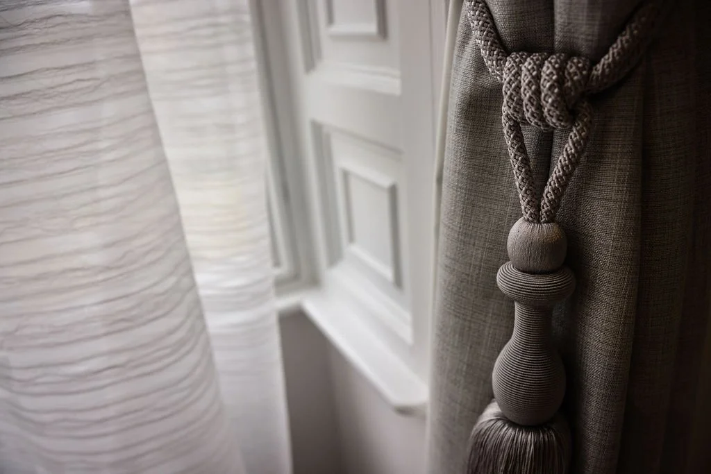 Close-up of a window with a white textured curtain and a gray curtain with a decorative tassel tieback.