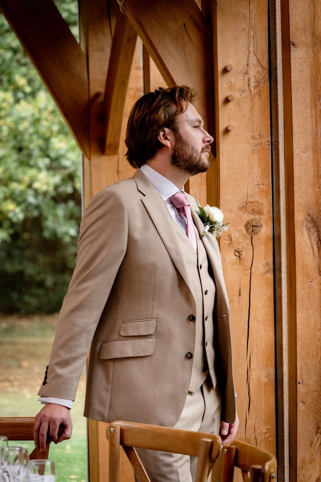 A man in a beige suit stands indoors, gazing out a wooden window, with a floral boutonniere on his lapel, in a setting with wooden walls and outdoor greenery.