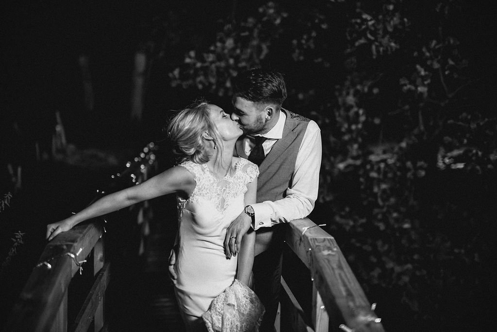 A black and white photo of a couple kissing on a bridge at night, with trees in the background.