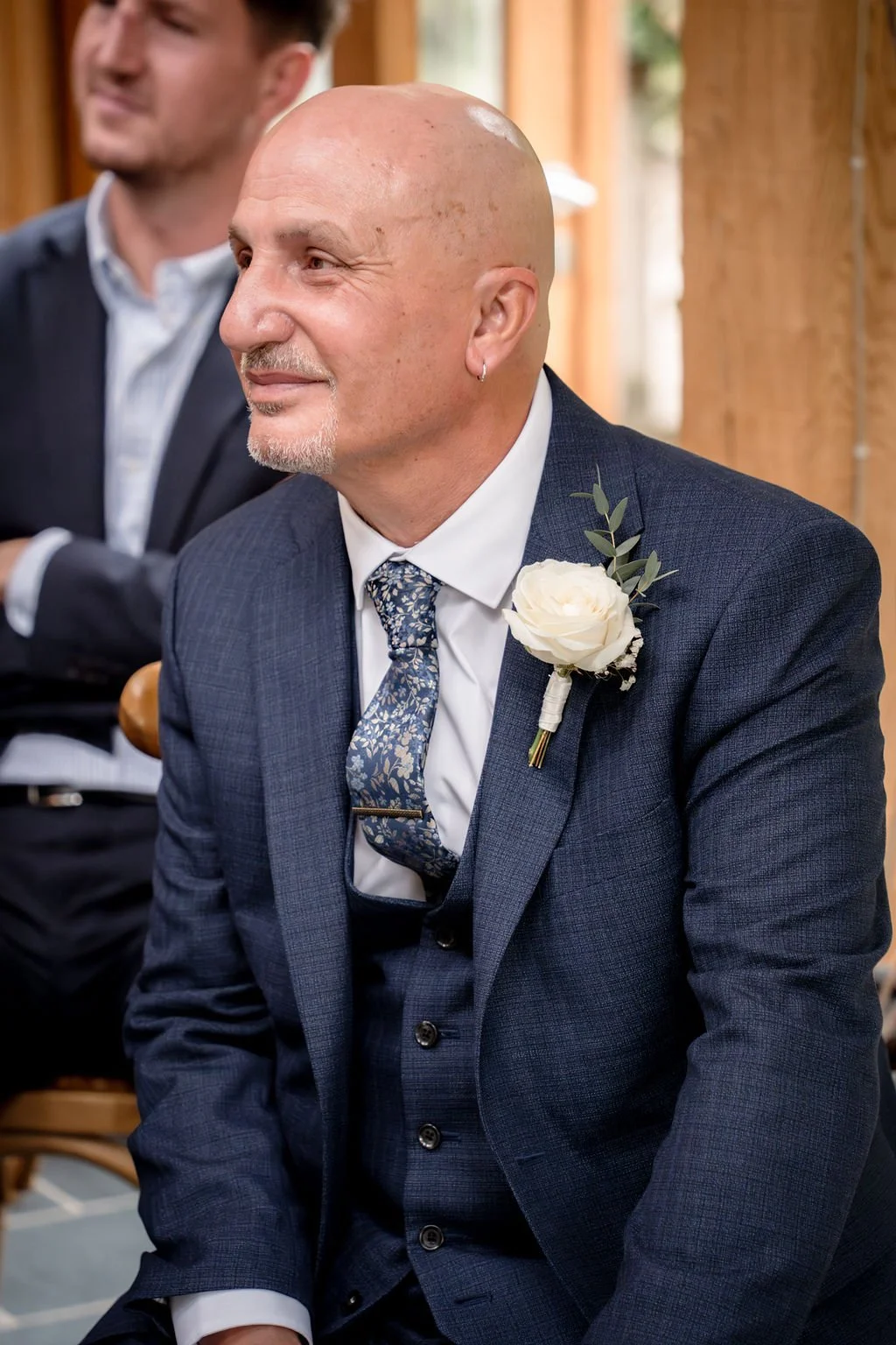 A bald man dressed in a navy suit with a floral tie and a white shirt, wearing a white flower boutonniere with greenery on his suit lapel, sitting and smiling at a formal event.