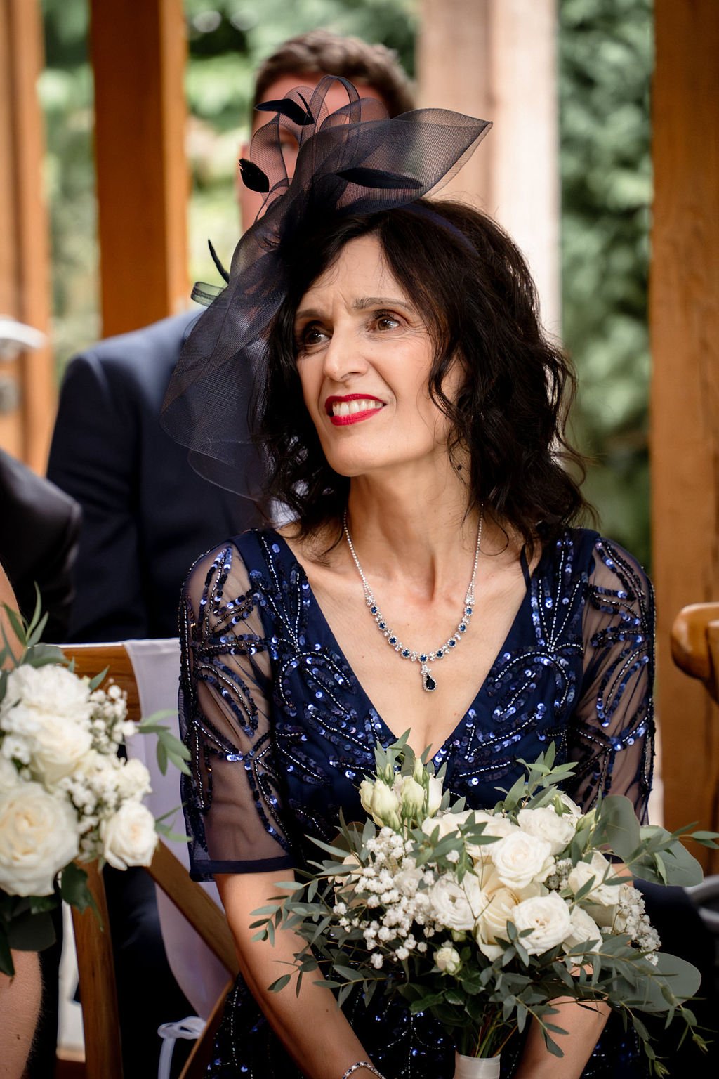 A woman with dark curly hair wearing a blue sequined dress and a black veil headpiece, holding a bouquet of white flowers during a wedding ceremony.