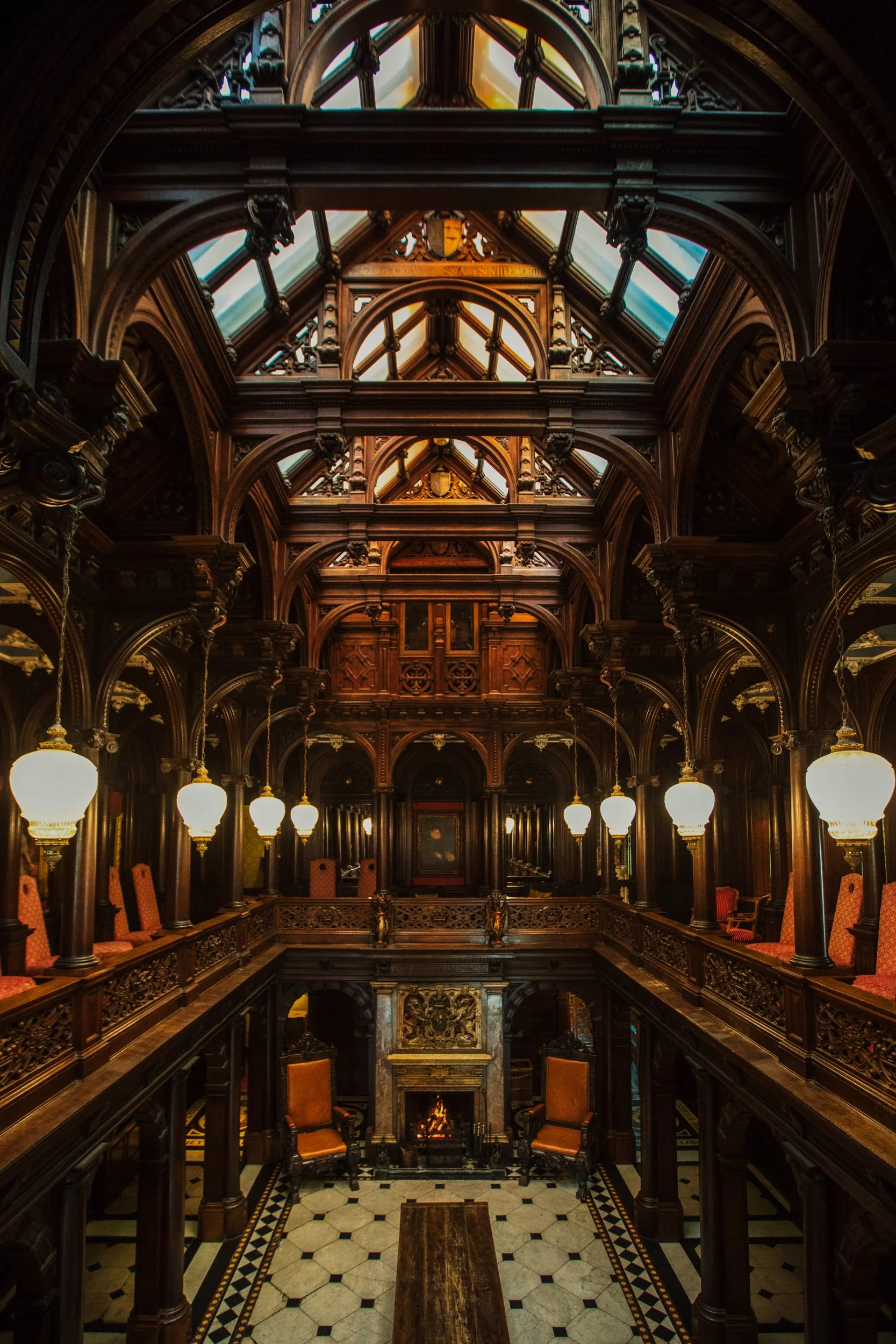 Interior view of a historic, elaborately decorated wooden building with a high, glass ceiling, ornate woodwork, hanging lamps, and furniture arranged around a fireplace.