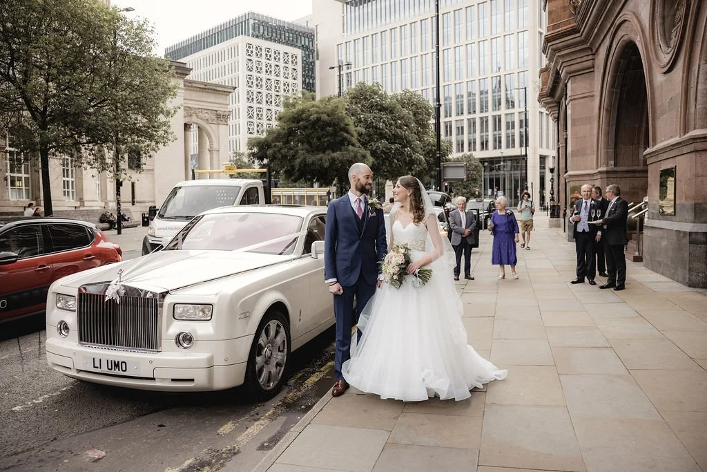 Bride and groom standing beside a white luxury car on a city street, with wedding guests in the background.
