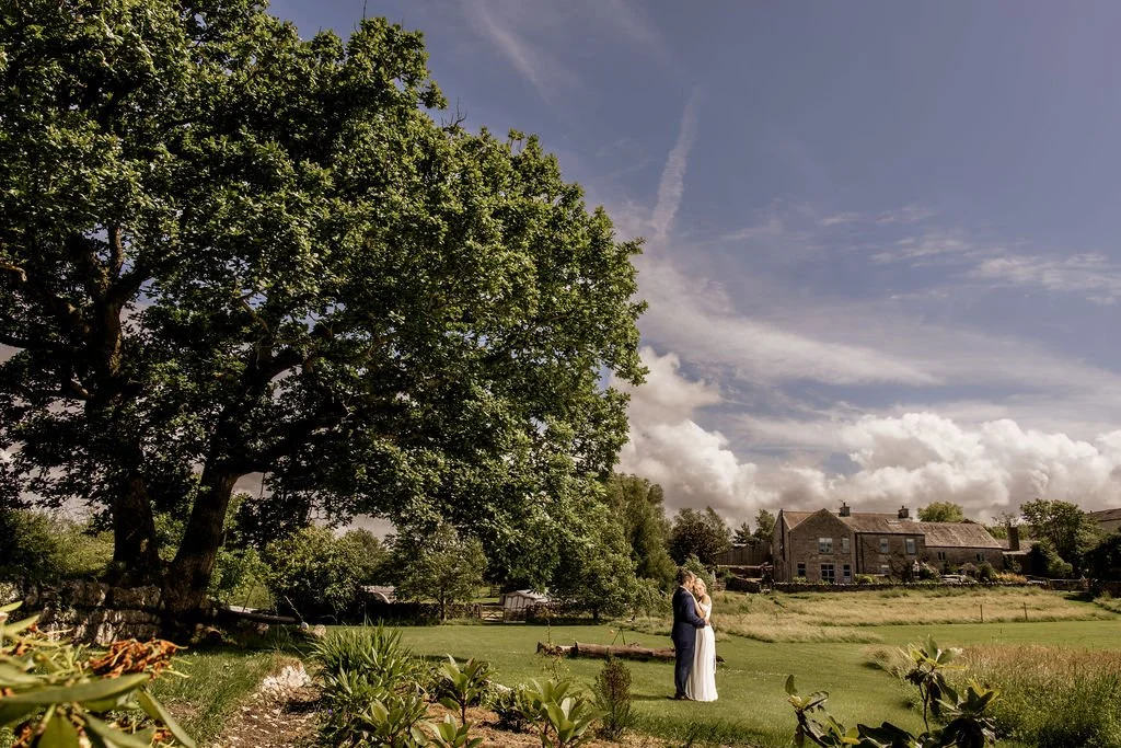 A bride and groom in wedding attire standing close together in a grassy field, embracing under a large tree with lush green leaves, with a rustic house and cloudy sky in the background.