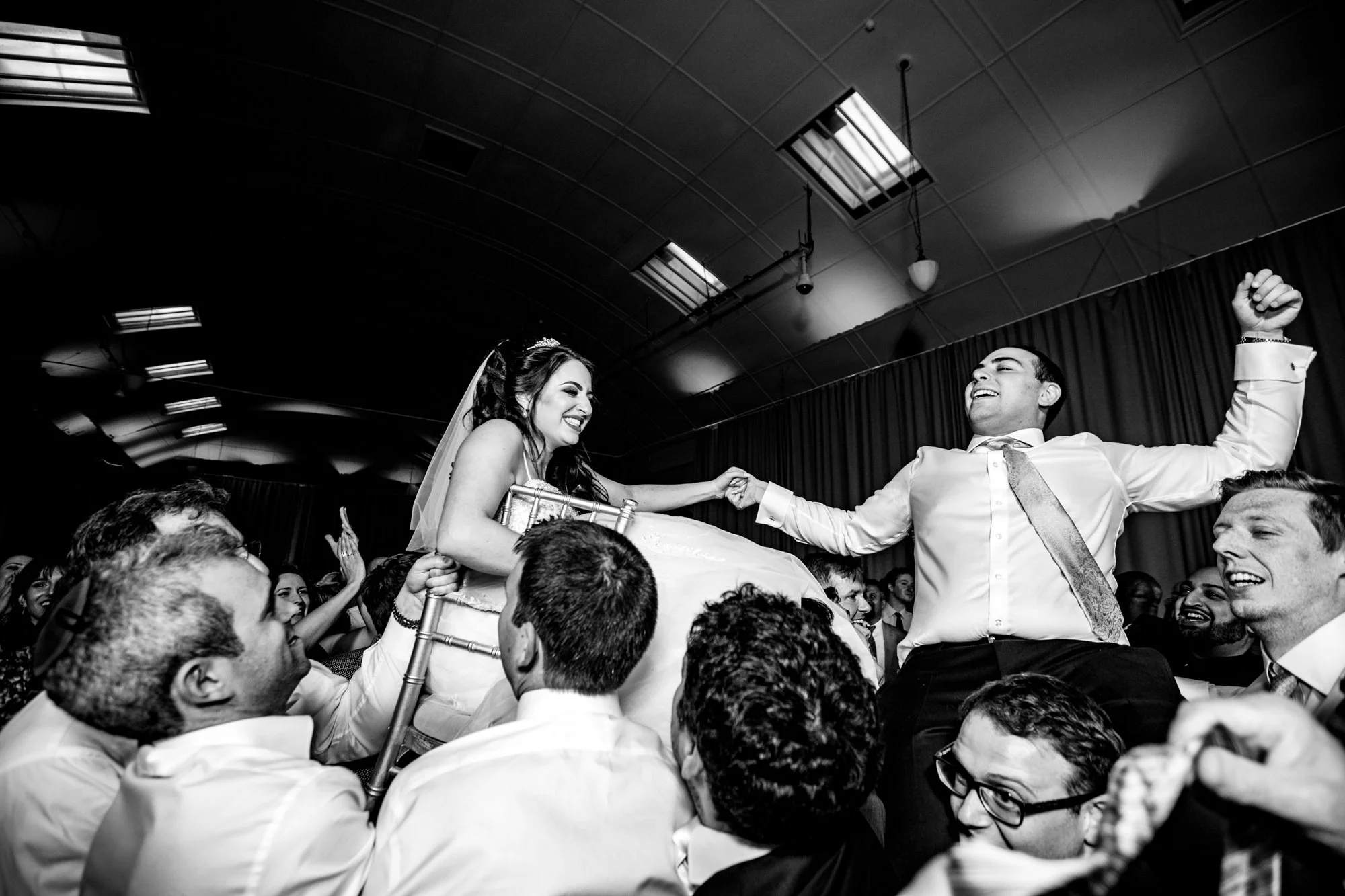 A bride and groom on a wedding cake during a celebration, surrounded by friends lifting the bride and groom, who are smiling and holding hands. The scene is lively and joyous.